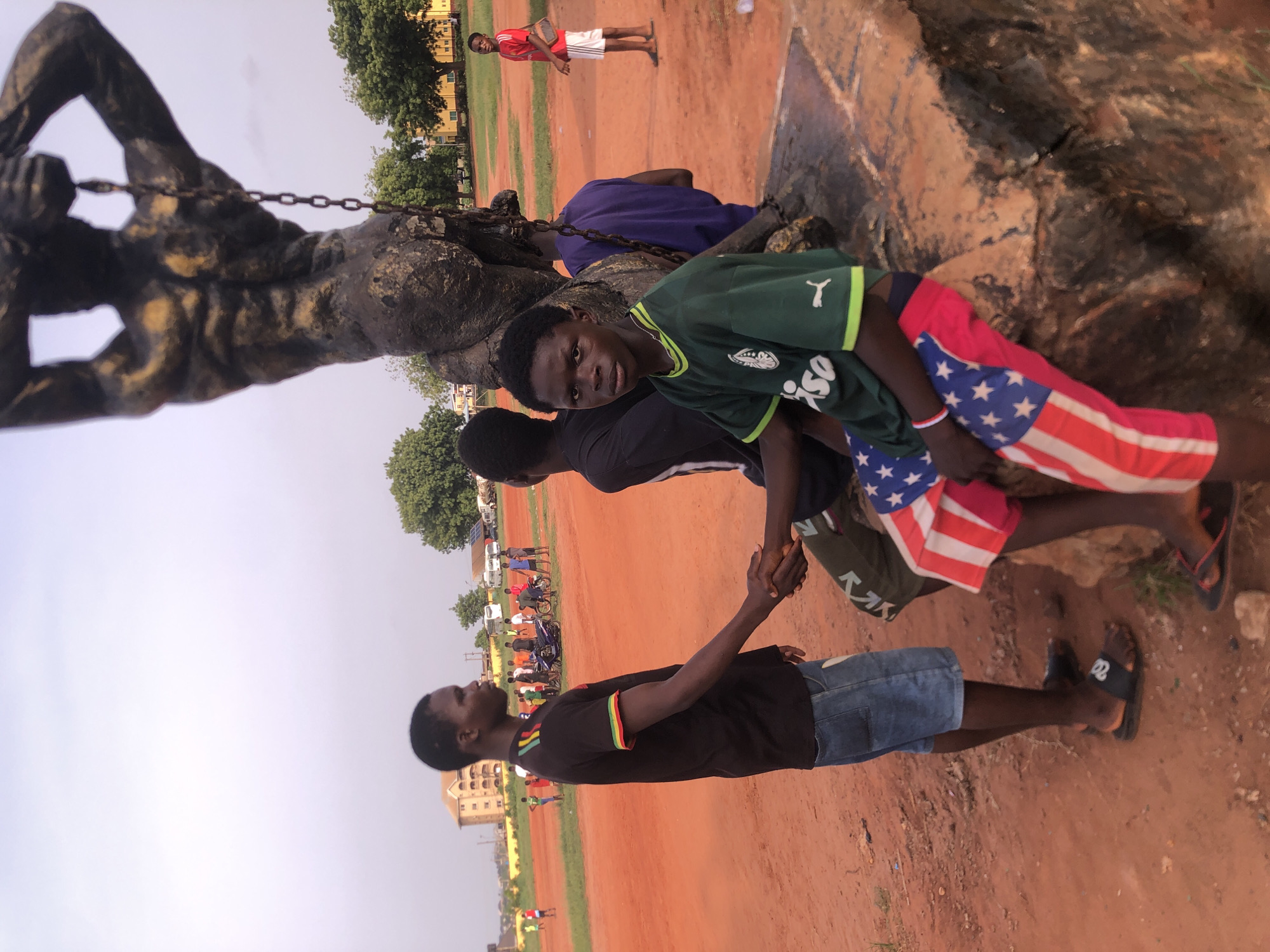 The image, rotated clockwise, depicts an outdoor scene in Ukwu-Abwa, Nigeria, featuring a large statue and several young men under a bright sky. The ground is reddish-brown earth, indicative of a dry, open field or public space.
In the left foreground, a dark, muscular statue of a man with chains around his wrists is prominent. Several young men are gathered around its base. One young man, wearing a green football jersey with a "Puma" logo and "Ciso" text, along with red, white, and blue shorts patterned like the US flag, looks directly at the camera. He has a red and white bracelet on his wrist. Beside him, another young man in a dark t-shirt and grey/camouflage shorts is shaking hands with a third young man. This third individual wears a black t-shirt with red, yellow, and green striped sleeves, denim shorts, and black slide sandals.
Further back, a young man in a purple t-shirt leans against the statue, partially obscured. In the mid-ground, a young man in a red t-shirt and white shorts stands alone on the expansive, dusty field. The background shows scattered green trees, some distant buildings, and a clear, bright sky, suggesting it is daytime. The primary activity is a handshake between two of the young men, while others observe or are positioned nearby. The overall scene captures a casual gathering in an outdoor public area.