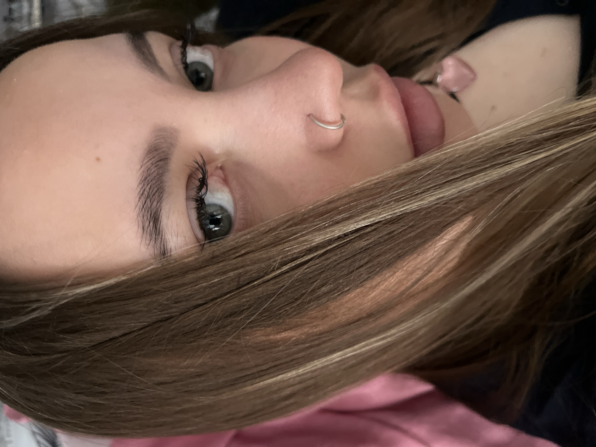 A close-up, slightly angled, indoor shot shows a young woman's face with brown and blonde hair partially obscuring her. She has green eyes with long eyelashes, light skin with a mole on her forehead, and a silver nose ring. Her lips are slightly parted. A pink, heart-shaped earring is visible near her left ear. The background is blurry and shows a pink and dark blue surface. The location of Novopokrovskaya, Russia is not evident from the image.