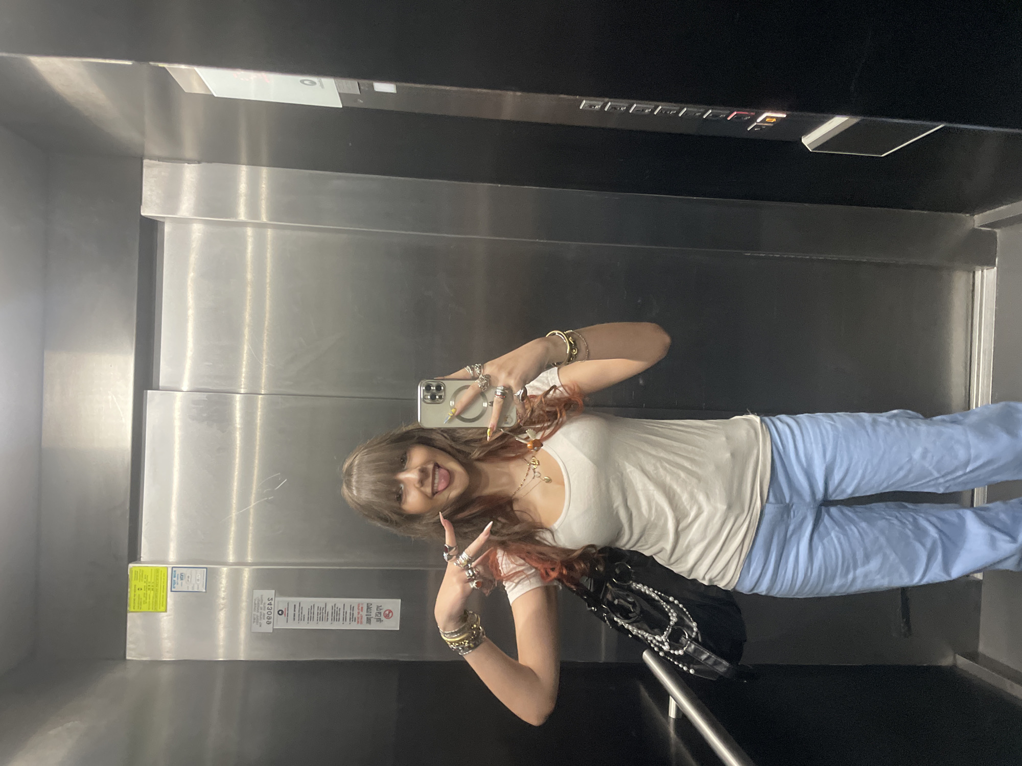 A young woman is standing in an elevator. She is wearing a white t-shirt and light blue pants. She has long brown hair with reddish highlights and is wearing several rings and bracelets. She is holding a smartphone in her right hand, taking a selfie. The elevator appears to be made of stainless steel and has a dark door on the right side with elevator buttons. There are some notices on the elevator wall. The scene is consistent with an elevator in an urban environment, potentially a residential or commercial building. Without additional context, it is not possible to definitively place the location in Raanana, Israel.