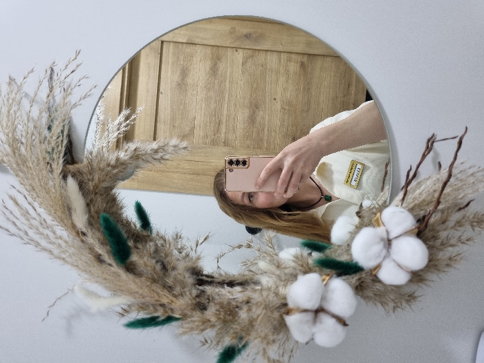 A circular mirror, partially obscured by a decorative arrangement, reflects a female subject capturing a selfie. The decorative arrangement around the mirror consists of dried pampas grass, dark green textured accents, and two prominent white cotton bolls. The reflected female has light brown hair and is seen from the chest up, holding a light pink smartphone with her right hand to photograph her reflection. She wears a light-colored collared shirt, a dark necklace with a green pendant, and a name tag that reads "Ольга" in Cyrillic script. The reflected background behind her is a light-toned wooden door or wall panel. The wall surrounding the mirror is a plain light gray. This scene is located in the city of Chaikovsky, Russia.