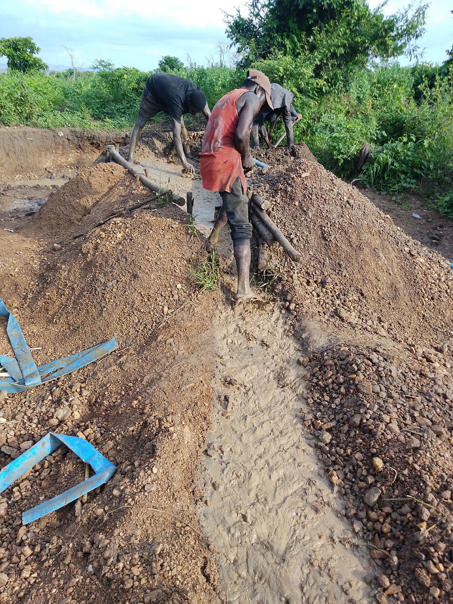 In Otulu, Nigeria, three individuals are engaged in manual labor at a muddy excavation site. Two men, dressed in dark clothing and one in an orange vest and dark pants, are actively working. One individual, wearing a black shirt and shorts, is bent over in the mud, appearing to dig with his hands or a tool. Another man, in an orange vest and dark pants, stands further back, also seemingly involved in the work. A third person, partially obscured and wearing dark clothing, is seen further in the background. The ground is a mix of dirt, mud, and gravel, with piles of excavated material. Blue plastic sheeting or piping is visible on the ground, likely used for drainage or containment. The environment is lush with green vegetation surrounding the site, suggesting a rural or undeveloped area. The sky is partly cloudy. The scene depicts a labor-intensive process, possibly related to construction or resource extraction, in a wet and earthy environment.