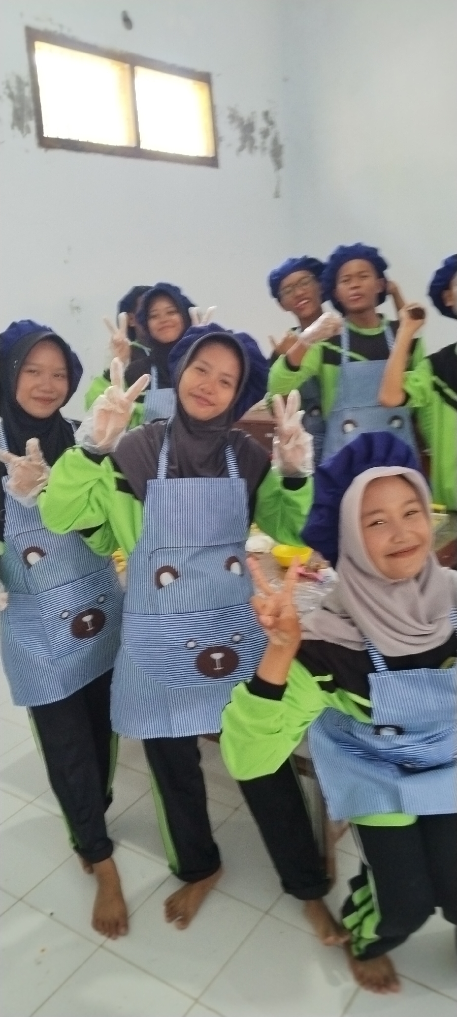 A group of nine young Indonesian individuals, appearing to be students, are posing for a photograph. They are all wearing blue chef hats and blue and white striped aprons with a teddy bear design. Most of them are also wearing light green long-sleeved shirts and black pants. Several individuals are wearing clear plastic gloves. Six of the individuals are in the foreground, with the remaining three behind them. All of them are making the peace sign with their fingers and smiling at the camera. The background appears to be an interior room with white walls and a window at the top left, possibly in a school or community center. The flooring is white tile. The overall impression is a cheerful and communal activity. Jombang, Indonesia, is a city known for its Islamic boarding schools, and this scene could be related to a cooking class or culinary activity at such an institution.