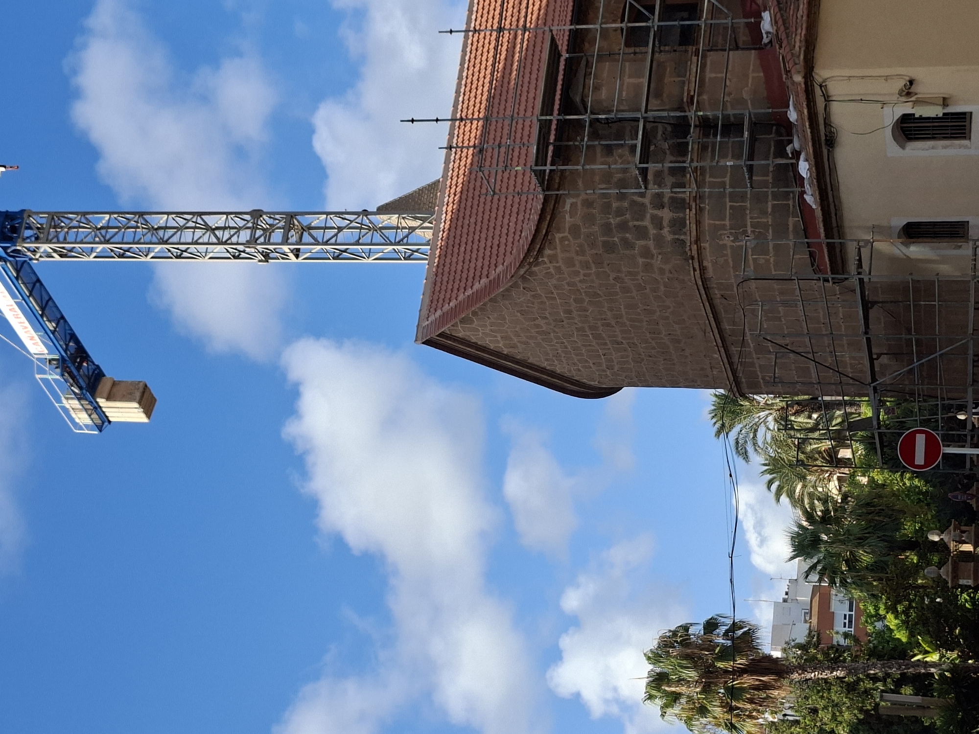 This image depicts a construction site in Torrevieja, Spain, with a tall blue crane dominating the upper portion of the frame against a bright blue sky filled with white clouds. In the foreground, a section of a building with a red tiled roof and stone walls is visible, partially obscured by scaffolding. The building appears to be undergoing renovation or construction. To the left of the building, palm trees and other lush greenery are present, suggesting a warm climate. A red and white "no entry" sign is also visible among the foliage. In the background, other buildings and palm trees can be seen, indicating an urban or suburban setting. The overall scene captures a moment of ongoing development within a tropical or Mediterranean environment.