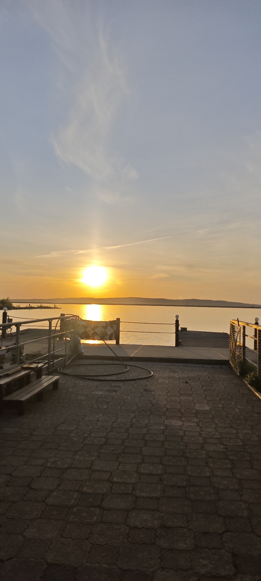 The image depicts a sunset over the Kama River in Chaikovsky, Russia. The sun is low on the horizon, reflecting brightly on the water. In the foreground, there is a paved area with a wooden bench and a metal railing. A coiled hose lies on the pavement. In the background, the opposite shore of the river is visible. The sky is a mix of blue and orange hues. No people are visible in the shot.