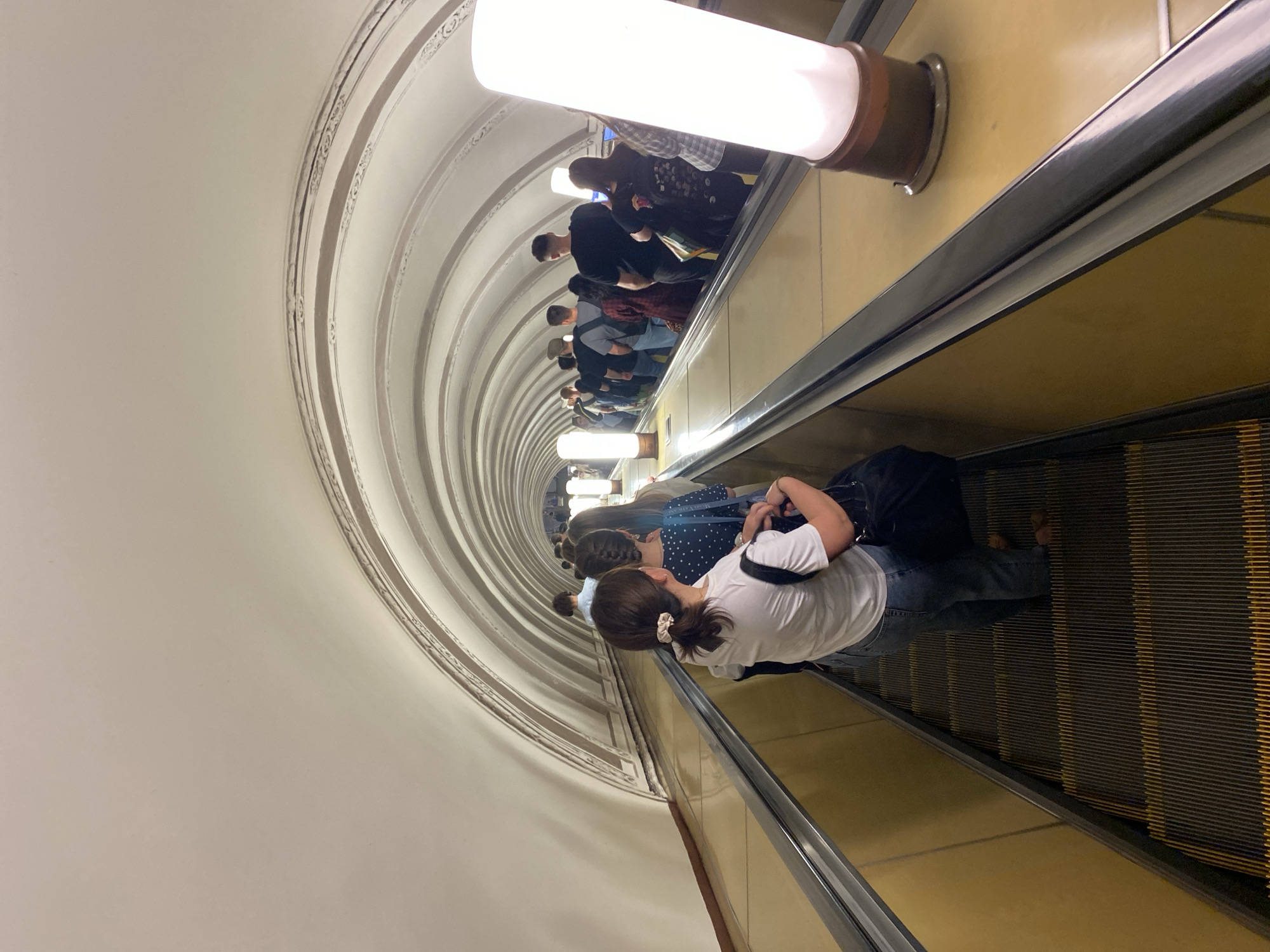 The image shows an escalator in the Moscow Metro. The escalator is descending into a tunnel lined with ornate, arched ceilings and lit by cylindrical lamps. A line of people, including a woman in a white shirt and jeans, are riding the escalator. The escalator steps are metal with yellow stripes. The walls flanking the escalator are gold.