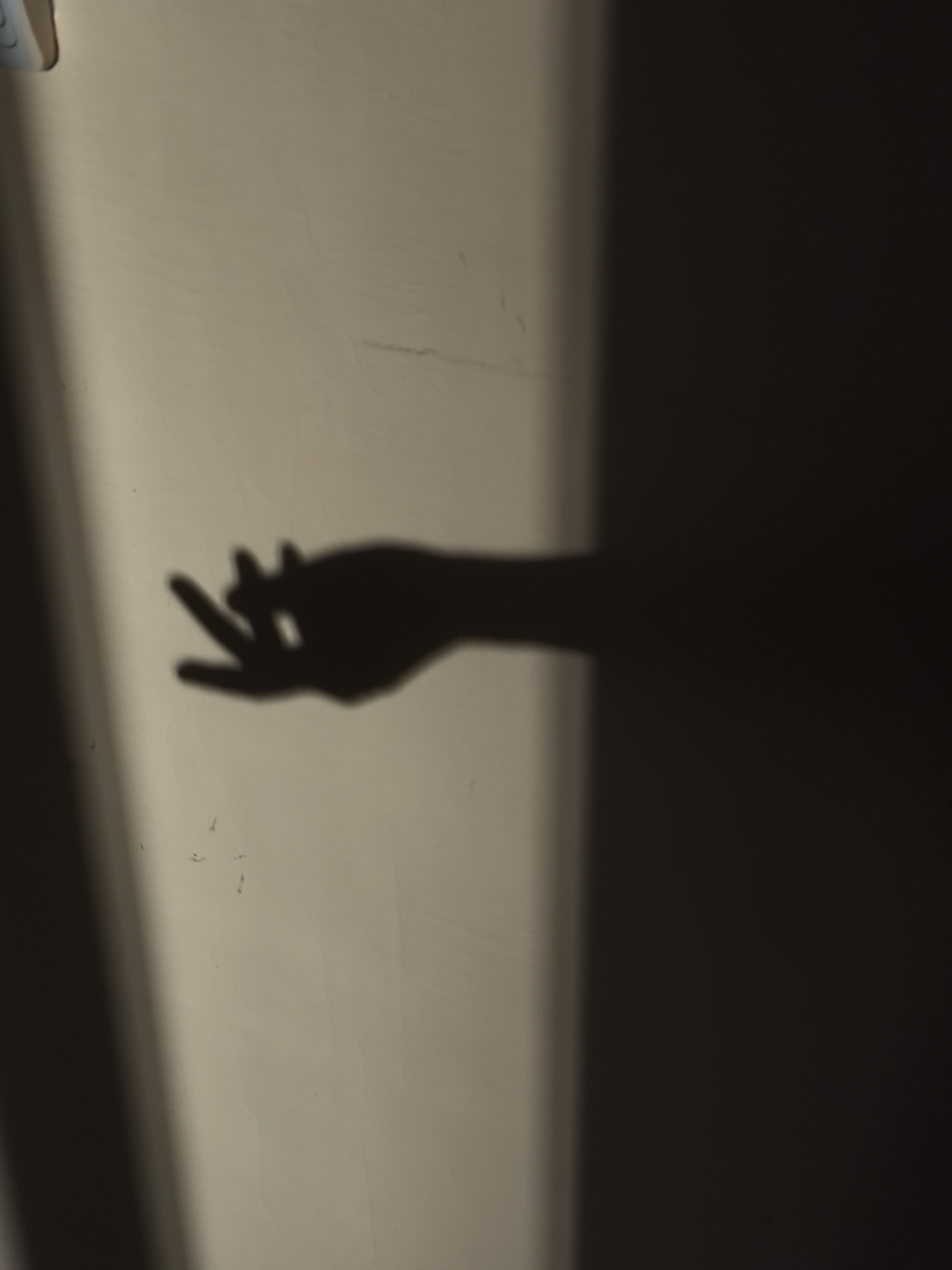 A shadow of a hand casts a peace sign against a wall. The hand appears to be reaching out to the left. The wall is textured and a pale beige color. A shadow from a vertical object is cast on the left side of the wall, creating a gradient of light and shadow. The right side of the image is dominated by a large dark shadow, possibly from the same object that casts the shadow on the left.