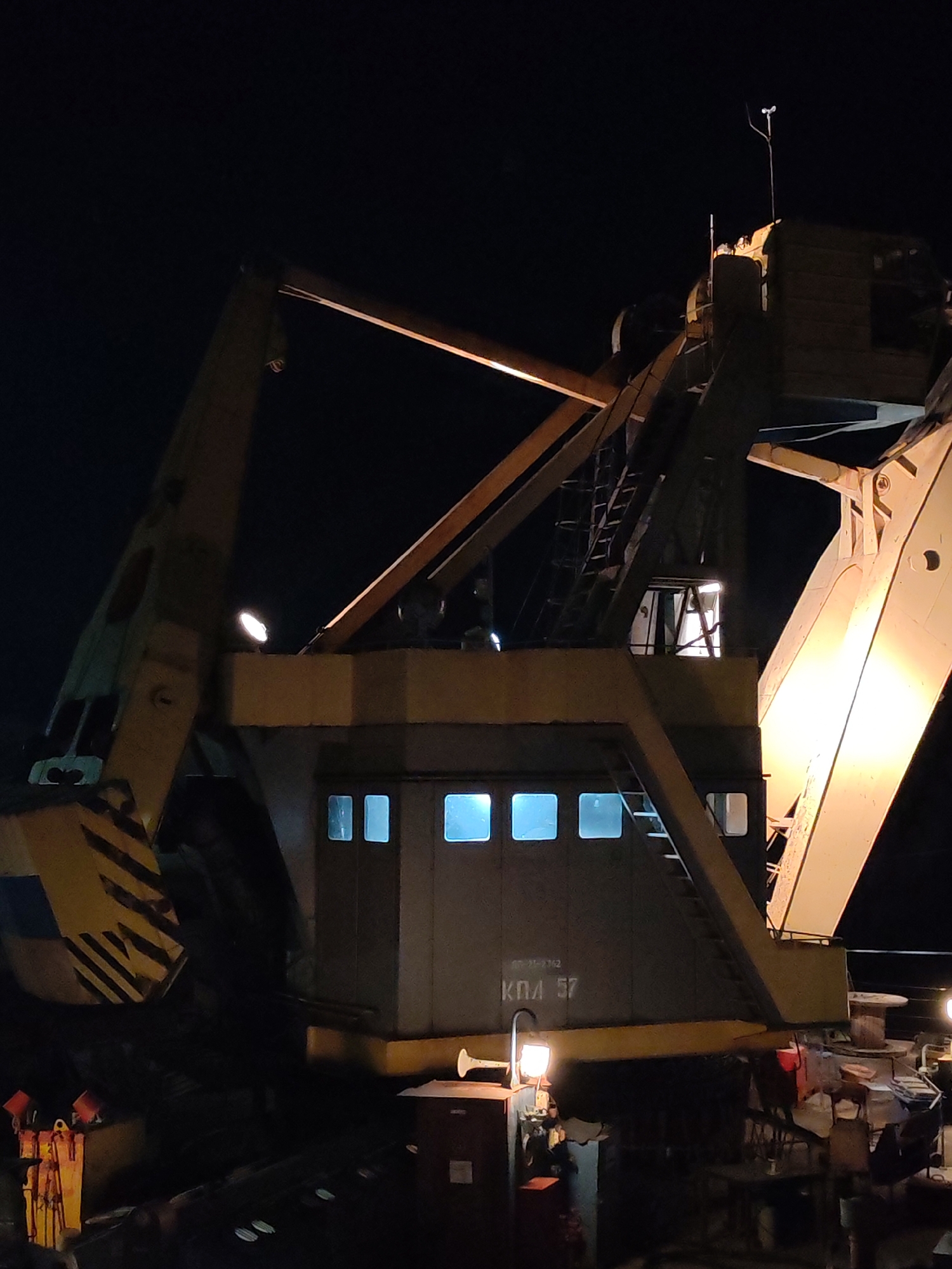 A medium shot at night reveals a large, industrial-looking, crane-like machine, likely used for construction or heavy lifting. The machine has a boxy, central structure with multiple small, square windows lit from within. The machine is painted in muted tones, possibly grey, with yellow accent stripes. On the side of the structure the Cyrillic inscription "KПЛ 57" can be seen. A complex framework of steel beams surrounds the box structure. A metal ladder is attached. A light source illuminates the area near the base of the structure, suggesting activity around the machine. The photograph was taken in Yakhroma, Russia.
