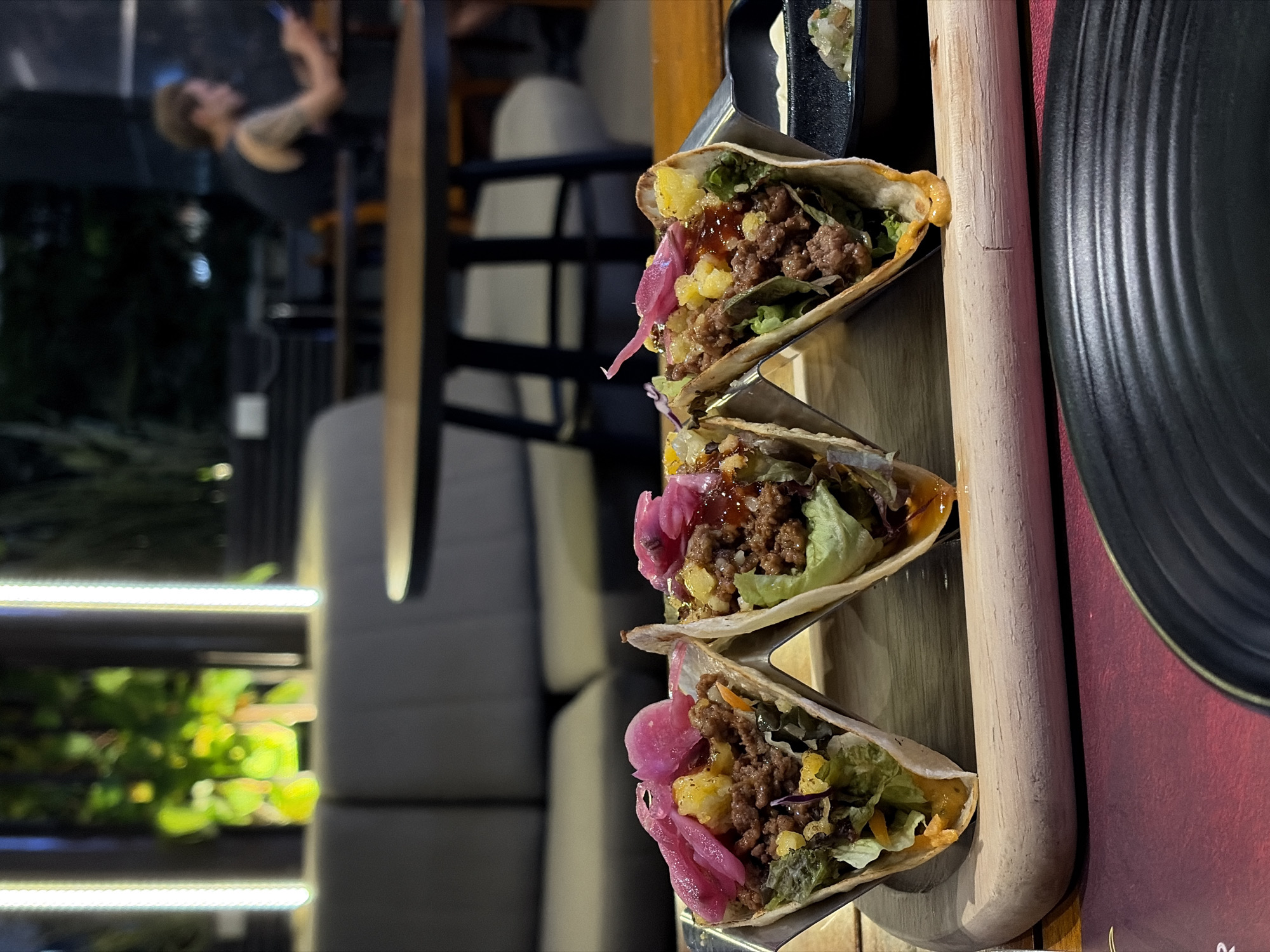 This media file features a close-up view of four tacos arranged vertically on a wooden serving board. The tacos are filled with ground meat, lettuce, and what appears to be pickled onions and corn. The background suggests an indoor setting, possibly a restaurant or bar, with blurred figures and seating visible. The lighting indicates it might be evening or nighttime, contributing to a warm and inviting atmosphere. In the far background, a person is visible, seemingly engaged with their phone. The presence of plants and illuminated lighting fixtures adds to the ambiance. To the right of the tacos, a black textured plate is partially visible, and a wooden pole runs vertically through the frame. The scene is set in Nha Trang Ward, Vietnam, hinting at a tropical or coastal location. No visible text is present in the media file.