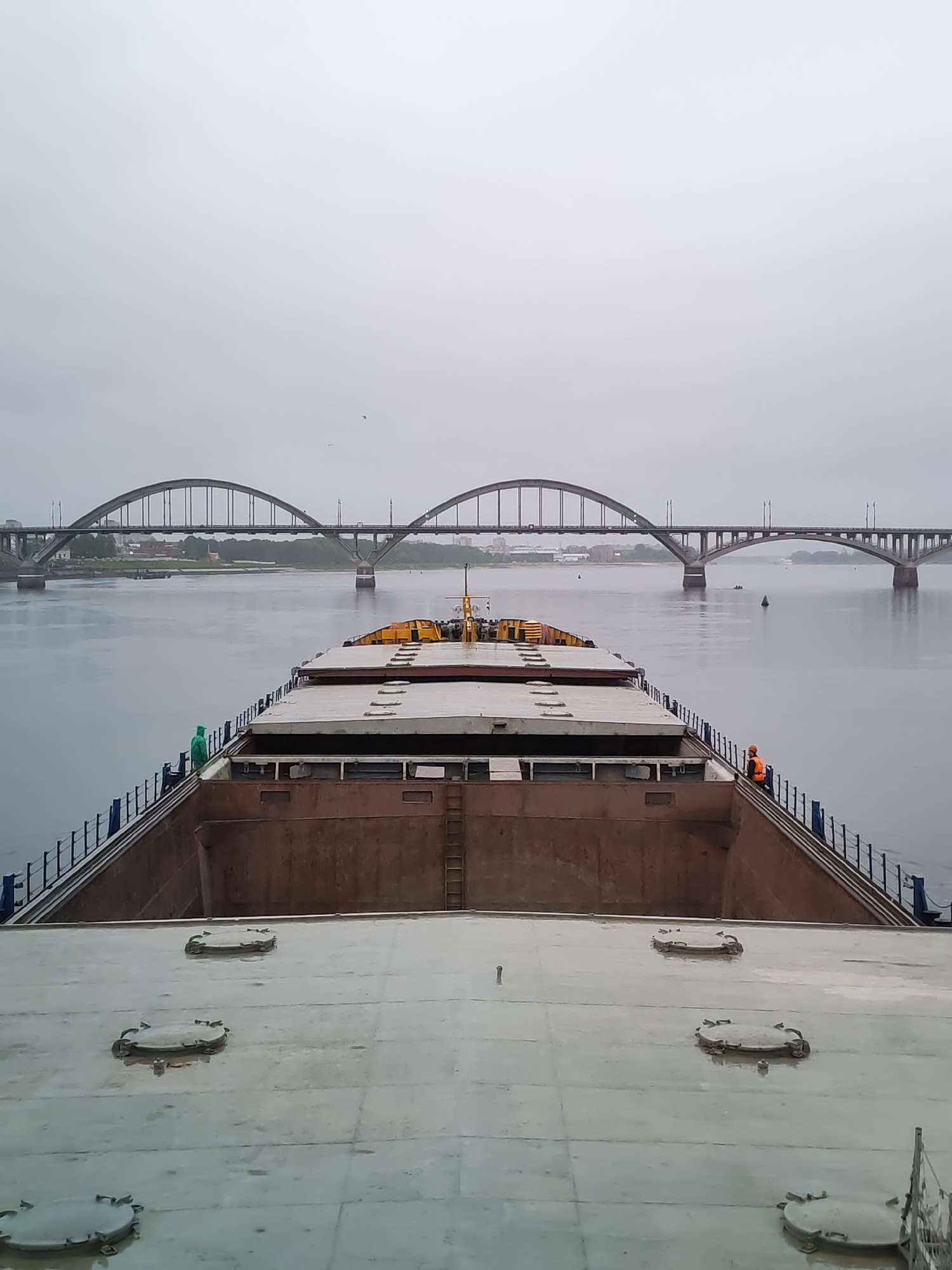 The image shows the deck of a large barge or ship, with a wide river and an arched bridge in the background. The sky is overcast. Two people wearing high-visibility vests are visible near the edge of the barge. There are several round hatches on the deck. The bridge appears to be the Arched Bridge over the Volga River in Bol'shie Mkhi, Russia.