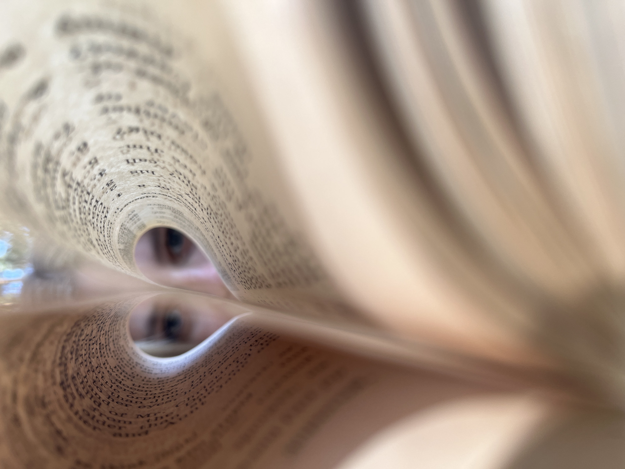 The image features a close-up perspective looking through a tunnel created by the curled pages of a book. The text on the pages is visible, but blurred. Two eyes are visible through the hole at the end of the tunnel. The lighting is soft and diffused, creating a warm tone. The book's pages appear to be off-white or cream-colored. The location in the city of Chaikovsky, Russia, is not determinable from this image.