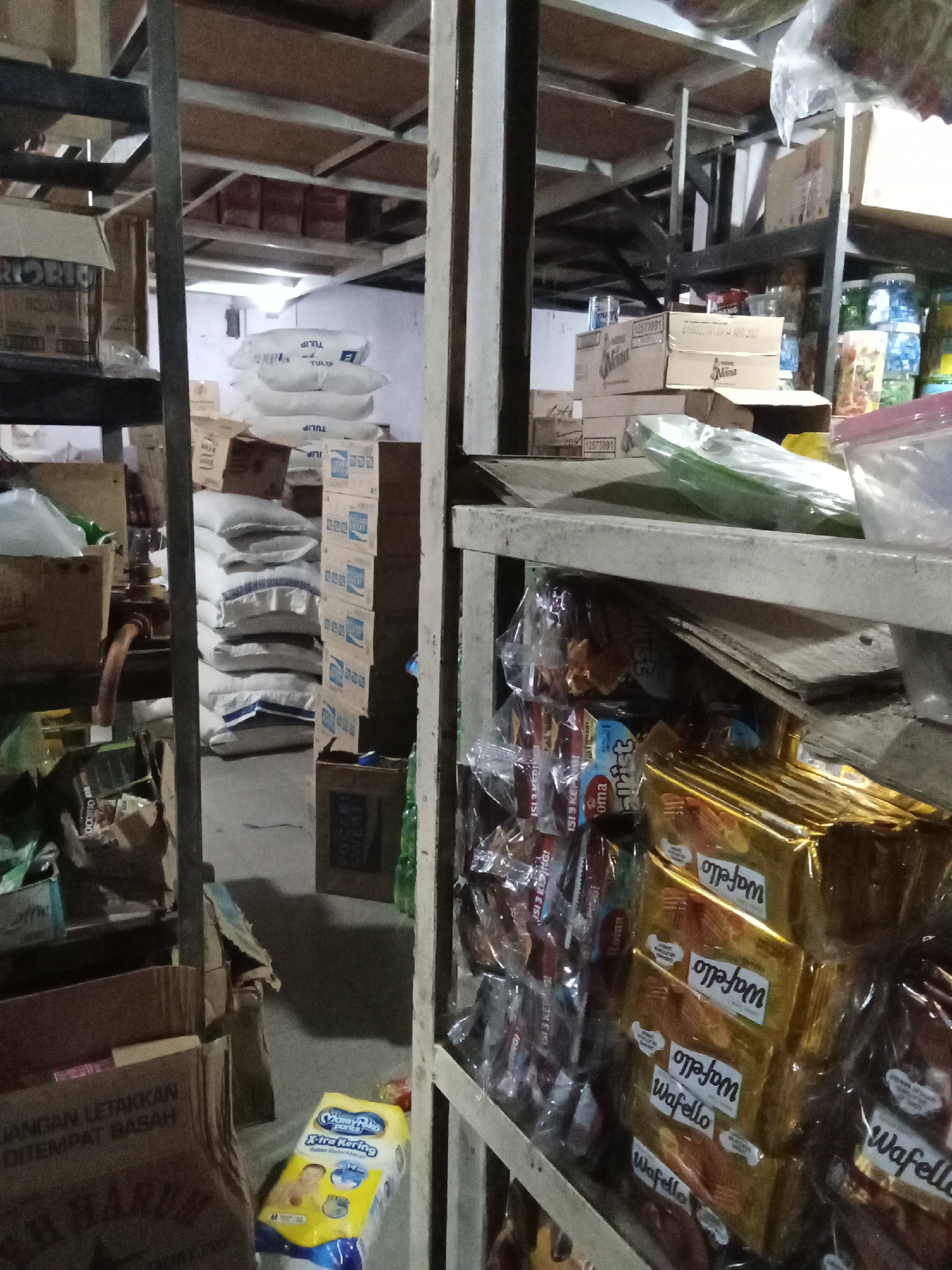 This is an indoor, medium-shot, eye-level photograph of a store. The image is cluttered with various products stacked on shelves and on the floor. In the foreground, there are several stacks of golden-wrapped snacks, possibly cookies or wafers, with "Wafello" printed on them. To the left of these snacks, there are rows of smaller packaged goods. On the floor in the lower left, there is a large brown cardboard box with Indonesian text on it, and to the right of that, a yellow package of baby diapers. In the background, there are stacks of white bags, possibly rice or flour, labeled "TULIP". Various cardboard boxes and shelves are visible throughout the scene. The lighting is dim, suggesting an interior space such as a storeroom or a small grocery store. The scene is consistent with the interior of a retail establishment in Ajamu, Indonesia, potentially a small warung or minimarket.