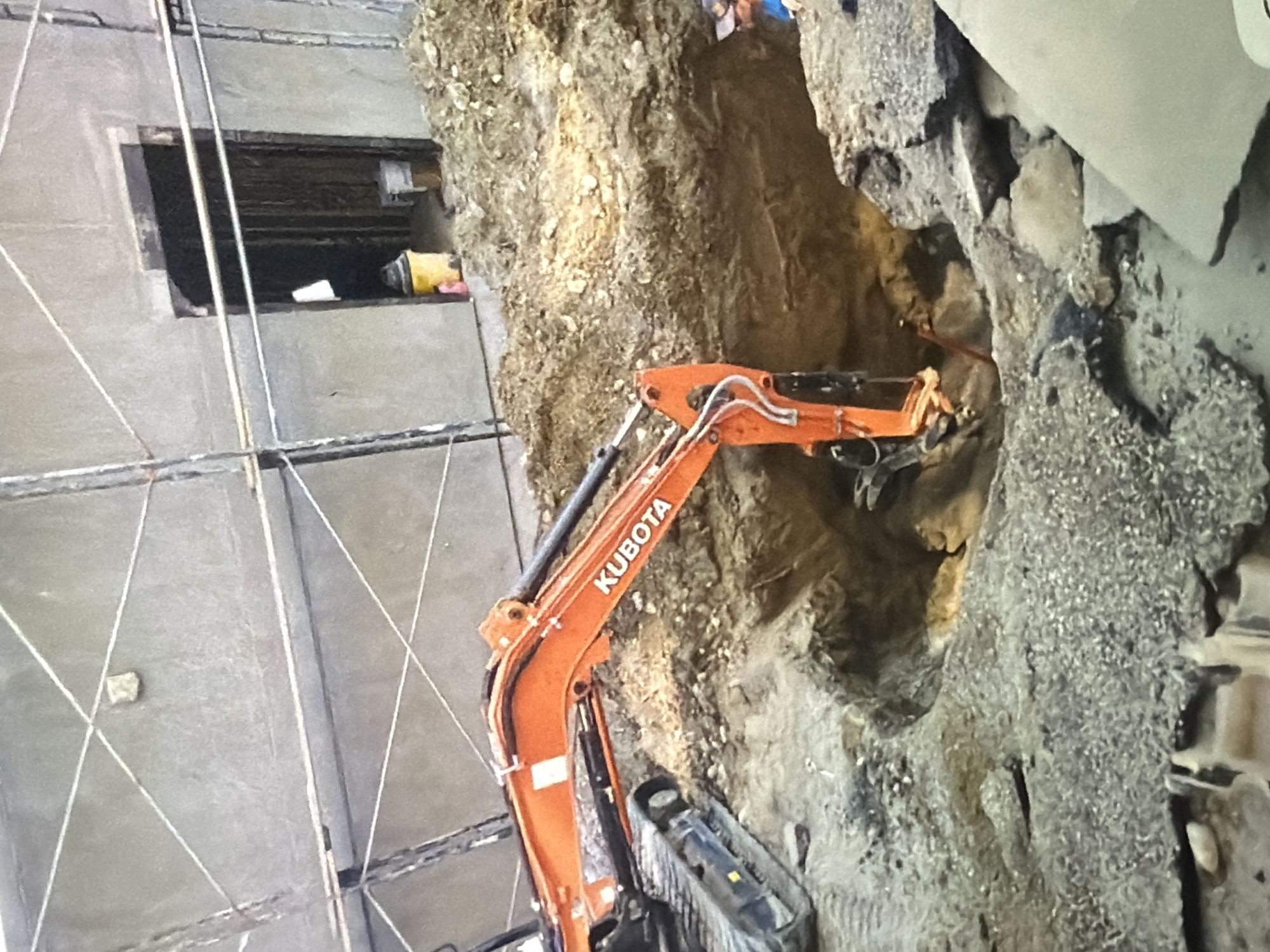 An orange Kubota excavator is positioned in a dirt excavation at a construction site, with a person in a blue shirt and a baseball cap observing from the right side of the frame. The excavation is deep, revealing layers of soil and rocks. In the background, a building under construction is visible, with scaffolding and a doorway leading into a dark interior. The ground is covered in dirt, gravel, and debris, with some concrete slabs present in the foreground. The scene suggests ongoing construction or demolition work.