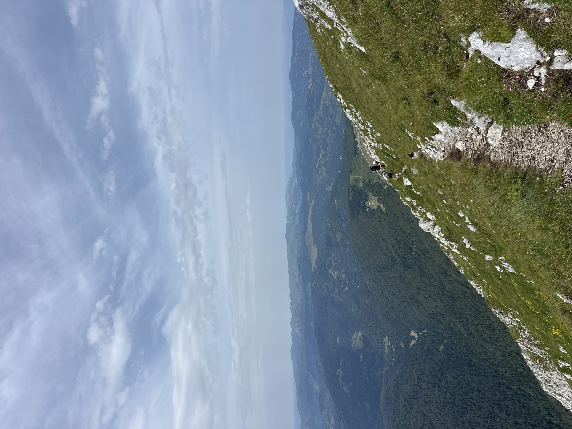 The image captures a scenic view from a mountain ridge in an unspecified location in Montenegro. In the foreground, a rocky, narrow hiking path winds along the steep, grassy slope. Several hikers, dressed in outdoor gear, are visible on the path, ascending the mountain. The path is dotted with white rocks and bordered by green grass and occasional yellow wildflowers. The middle ground features a vast expanse of rolling, forested mountains fading into a hazy horizon. The sky above is a mix of blue and white clouds, with wispy cirrus clouds streaking across the upper portion of the frame. The overall scene depicts a mountainous landscape with evidence of outdoor activity.