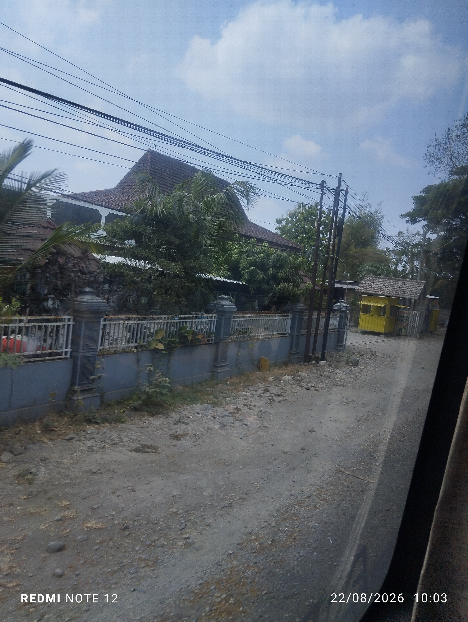 The image shows a street scene in Karangrejo, Indonesia. On the left, a house with a tiled roof and white railings is partially visible, surrounded by lush greenery and palm trees. A grey concrete wall topped with a white metal fence runs along the front of the property. A dirt road with scattered stones and dry grass occupies the foreground and extends to the right. Further down the road on the right, a small, bright yellow building with a tiled roof stands, possibly a stall or kiosk. Utility poles with numerous overhead wires are prominent in the scene. The sky is partly cloudy with a bright blue expanse. The bottom of the image contains text indicating "REDMI NOTE 12" and a date stamp "22/08/2026 10:03". No people are visible in the frame.
