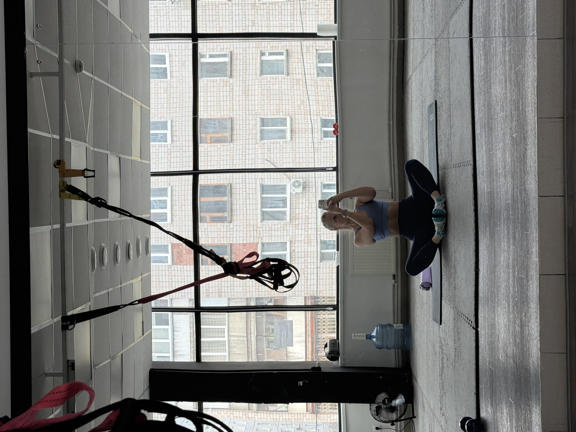 A woman sits on a mat in a gym, taking a photo with her phone. The gym features a wall of windows, through which an apartment building in Izhevsk, Russia, can be seen. Gym equipment, including suspension training straps, are visible. A water cooler and fan are also present.