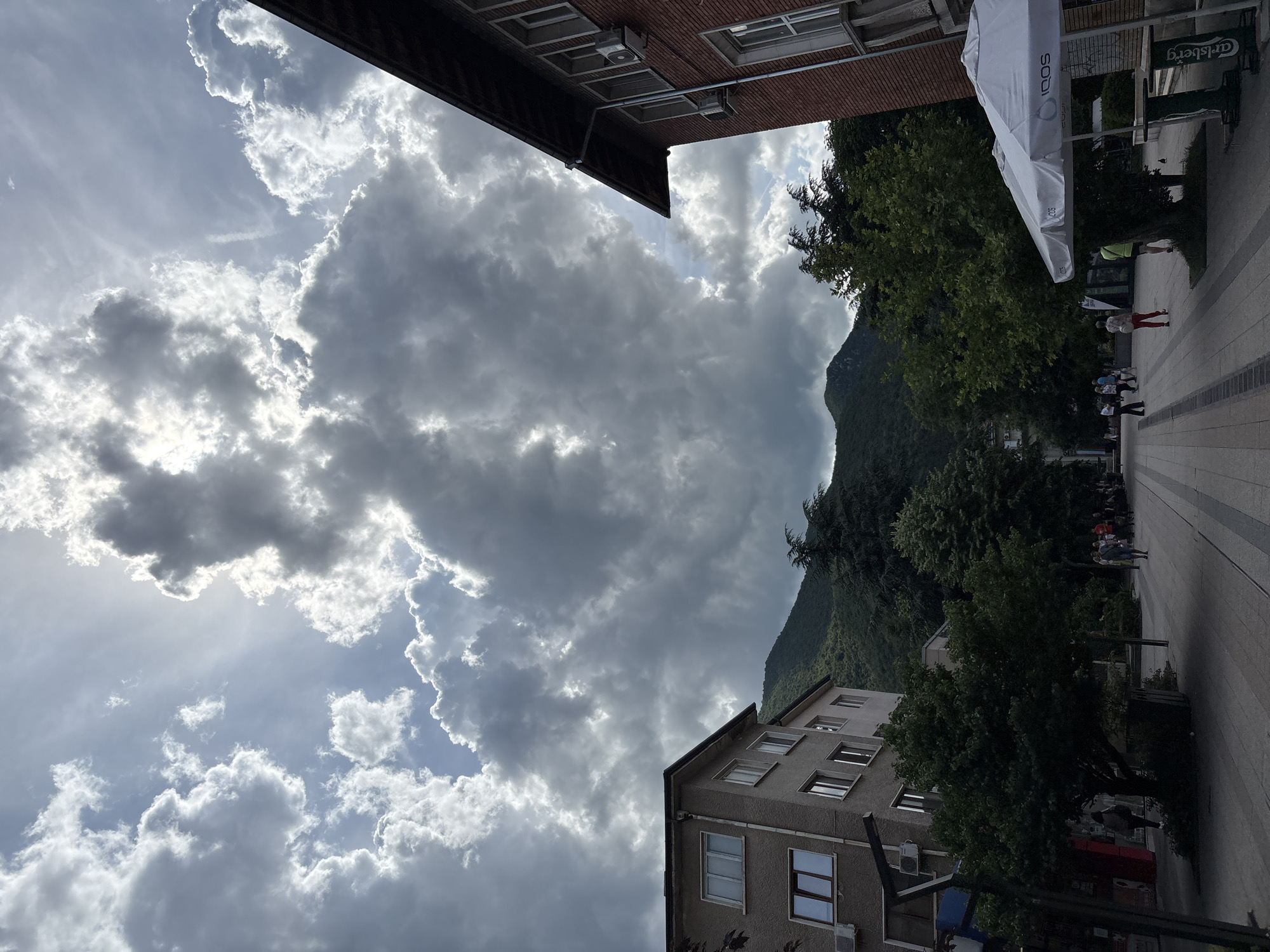 The image shows a street scene in Vratsa, Bulgaria. The sky is filled with dramatic, gray clouds, with the sun breaking through in some areas, creating a dramatic effect. In the foreground, on the right, is a red brick building with a covered outdoor seating area in front of it, complete with white umbrellas and tables. On the left, there is a lighter-colored building. The street is paved with patterned tiles and is lined with trees. Several people are walking along the street, some with their backs to the camera. In the background, a lush green mountain rises, partially obscured by trees and the cloudy sky. The scene suggests a busy urban environment with a natural backdrop.