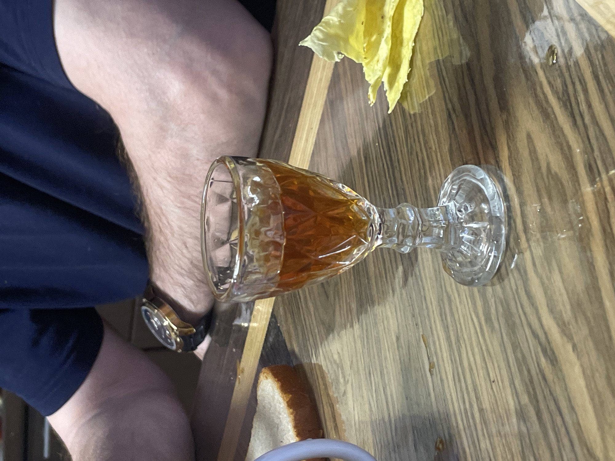 A high-angle shot depicts a table setting with a glass of amber-colored liquid, possibly liquor, laying on its side. The table has a light wood grain finish. A piece of bread and some yellow vegetable matter are also visible. A person is seated at the table, wearing a dark blue garment and a watch with a dark band. The location is unknown, and there is no information indicating the city of Kuyeda, Russia.