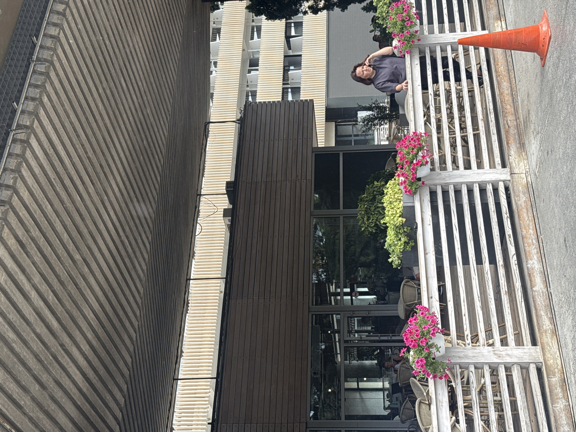 A woman is standing near a white wooden fence with pink flowers, in what appears to be an outdoor seating area of a cafe or restaurant. She is talking on her mobile phone. Behind the fence, there are tables and chairs. In the background, there is a building with balconies. There is an orange traffic cone on the ground to the right of the fence. The scene is under a gray, ribbed overhang. The overall environment suggests an urban setting.