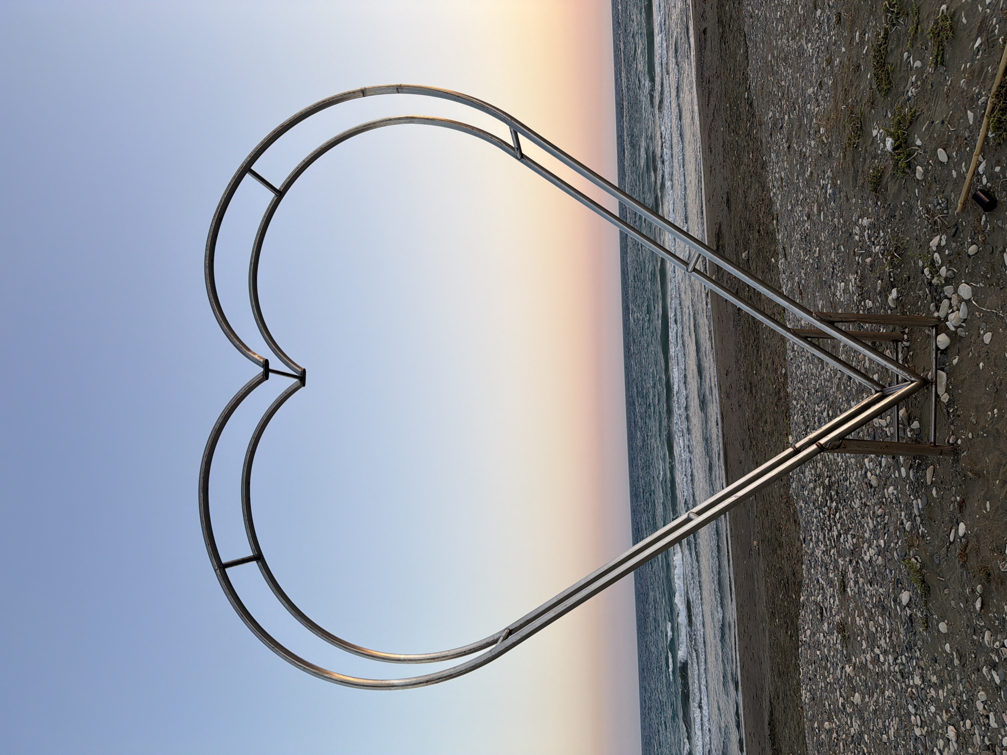 A large, metallic heart-shaped sculpture stands on a beach in Meneou, Cyprus. The sculpture is open, with two concentric heart outlines made of metal tubing, supported by angled legs. The beach is composed of sand and pebbles. Beyond the beach, the sea is visible with gentle waves breaking. The sky above is clear, transitioning from a pale blue at the top to soft hues of pink and orange near the horizon, indicating either sunrise or sunset. The scene is devoid of people.