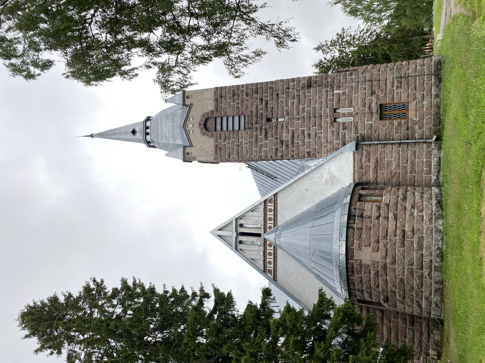 The image shows a stone church with a tall, pointed steeple, surrounded by trees. The church features a combination of light-colored stucco and rough-hewn stone construction. The steeple is made of gray metal and has several small windows. The lower portion of the church and the base of the steeple are constructed from large, reddish-brown stones. The church has several windows, some with lattice work. In the foreground, there is green grass. The sky is overcast. The provided information indicates the location is the city of Melnikovo, Russia.