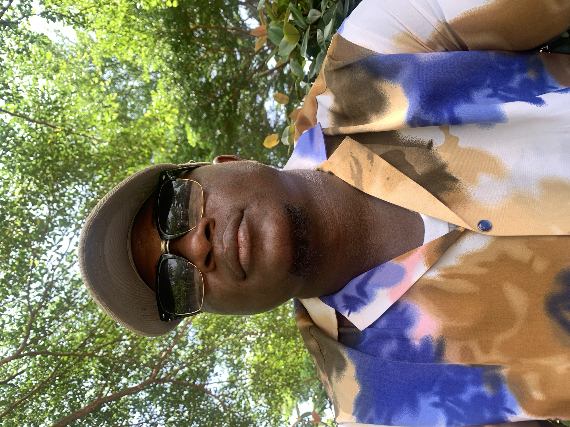 A medium shot captures a man from the chest up, wearing a tan baseball cap, sunglasses, and a colorful tie-dye shirt over a white t-shirt. He has a goatee and is looking directly at the camera. The background is a blur of lush green foliage and tree branches, suggesting an outdoor setting. The lighting indicates it is daytime. The man's location is identified as Akwanga, Nigeria.