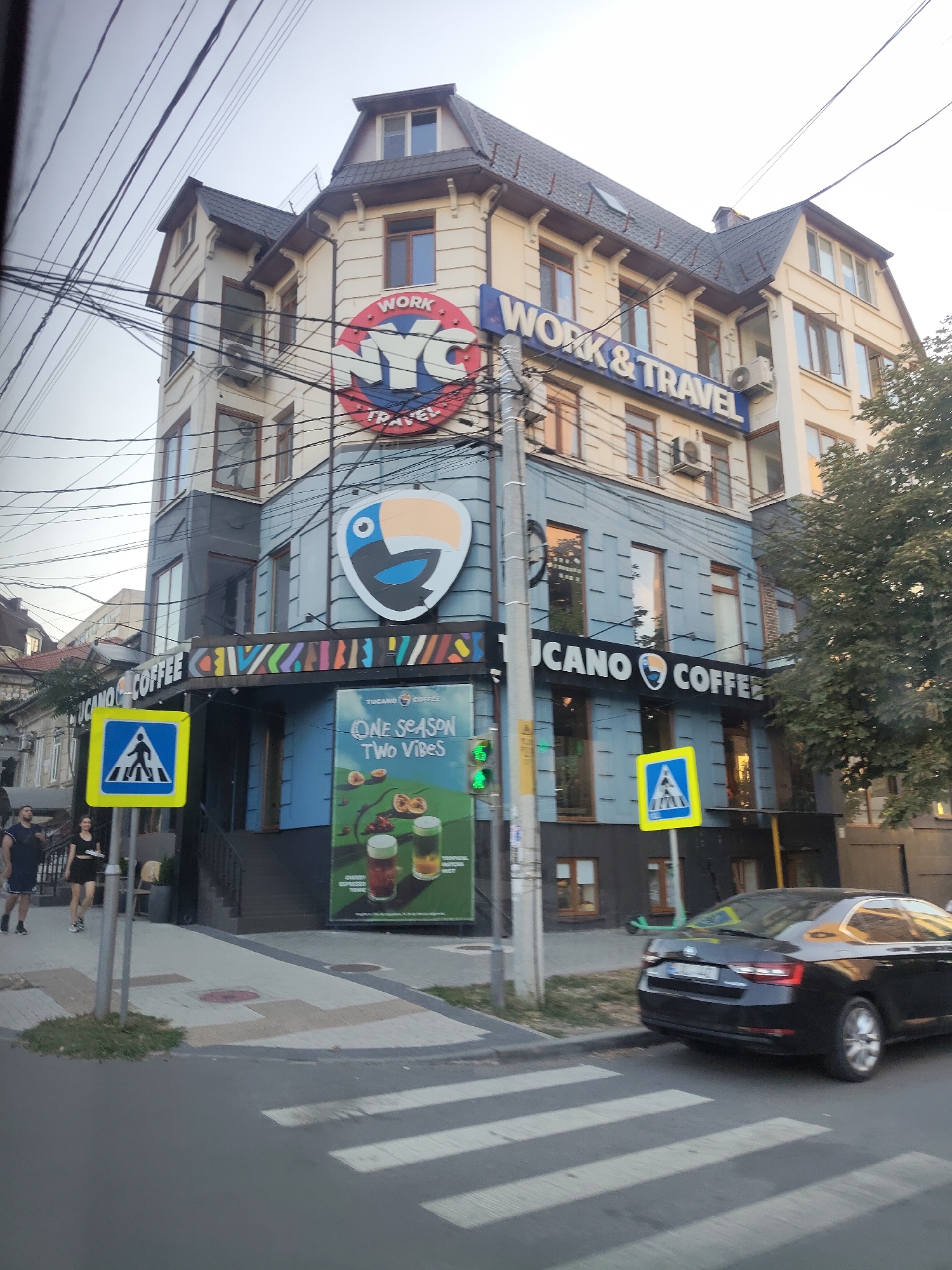 A street scene in Chișinău, Moldova, shows a multi-story building with businesses on the ground floor. The building has a beige facade with dark tiled roofs and numerous windows. The upper portion of the building features signage that reads "WORK & TRAVEL" and "NYC TRAVEL". Below this, the building's facade transitions to a blue-grey color, with a large graphic of a toucan. The ground floor is occupied by "TUCANO COFFEE", indicated by prominent signage. A pedestrian crossing is visible in the foreground. To the left of the building, two individuals are walking on the sidewalk, and a yellow pedestrian crossing sign is posted on a pole. A black car is parked near the pedestrian crossing. The sky is overcast, and power lines crisscross the scene overhead.