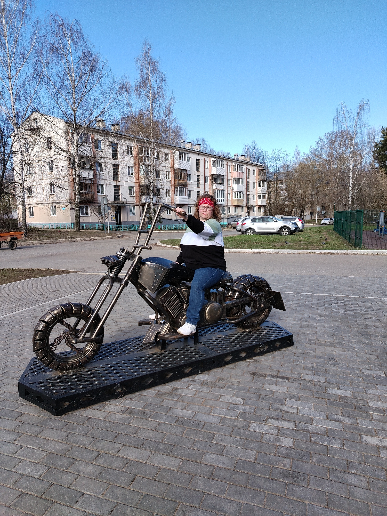 A young woman with glasses and a red bandana is sitting on a motorcycle sculpture in the city of Chaikovsky, Russia. She is wearing a black, white, and light green striped sweater, blue jeans, and white sneakers. She is pointing to the right. The motorcycle is a metal sculpture on a black metal platform. The ground is paved with grey rectangular bricks. In the background, there is a five-story apartment building, trees, a road, cars parked on the grass, and a green fence with a playground visible behind it. The sky is blue.