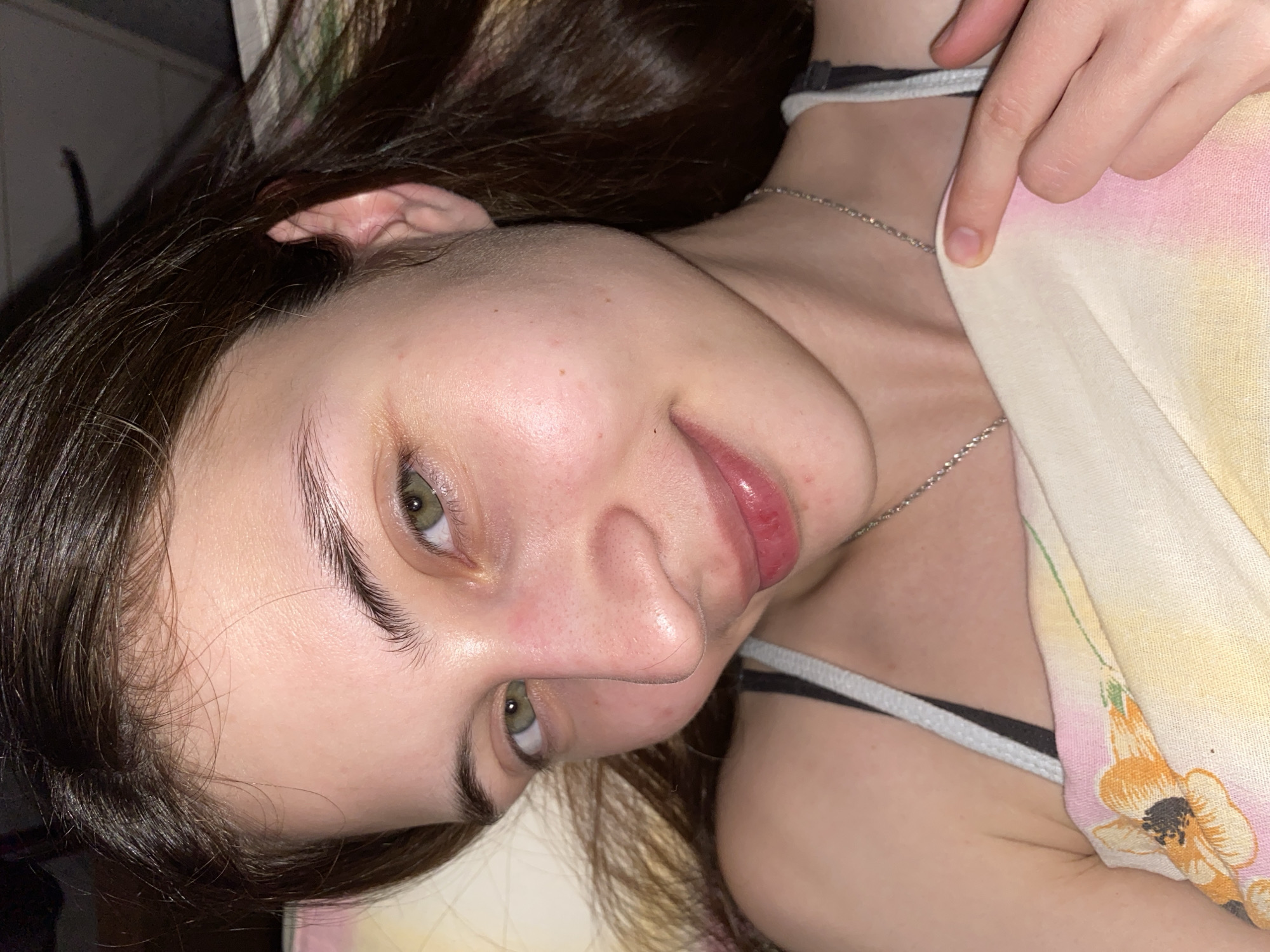 A close-up, tilted portrait of a young woman with long, dark brown hair. Her face is facing slightly upwards, with green eyes looking towards the viewer. She has fair skin with some minor blemishes. The lighting is soft and warm. She is wearing a patterned garment, possibly a camisole, with a black and white strap visible. A thin silver necklace rests on her collarbone. The background is indistinct and suggests an indoor setting. There is no indication of a location in the city of Votkinsk, Russia, or any identifiable objects or landmarks to suggest a specific geographic location.