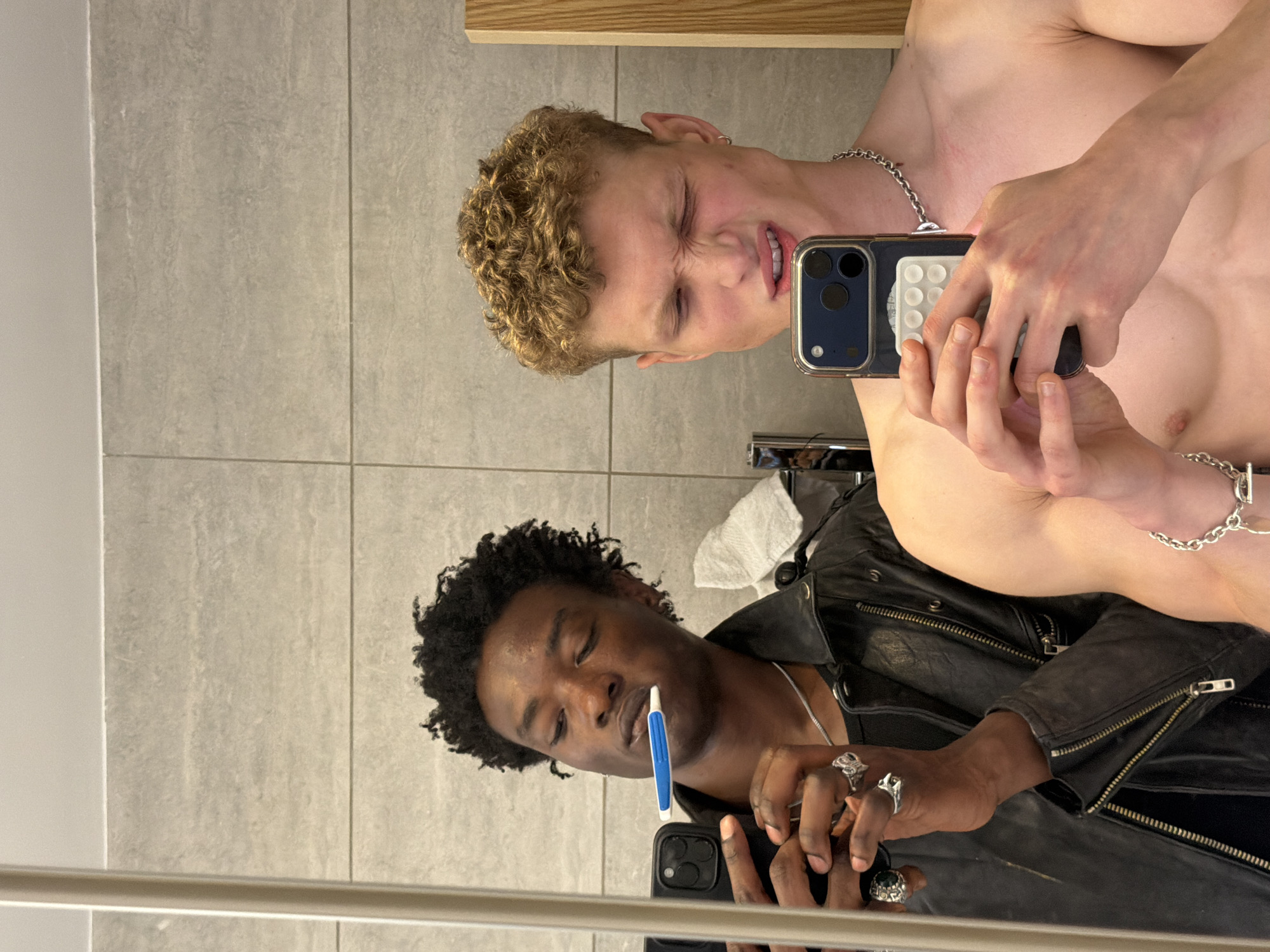 Two men are visible in a mirror reflection. The man on the top of the reflection is a shirtless white man with curly blonde hair. He is holding a phone up to his face and his eyes are shut tight. The man on the bottom of the reflection is a Black man with short, dark, curly hair. He is wearing a black leather jacket and holding a blue and white electronic cigarette in his mouth and a phone in his hand. He is also wearing rings on his fingers. The background is a grey tiled wall. The location cannot be determined from the image.