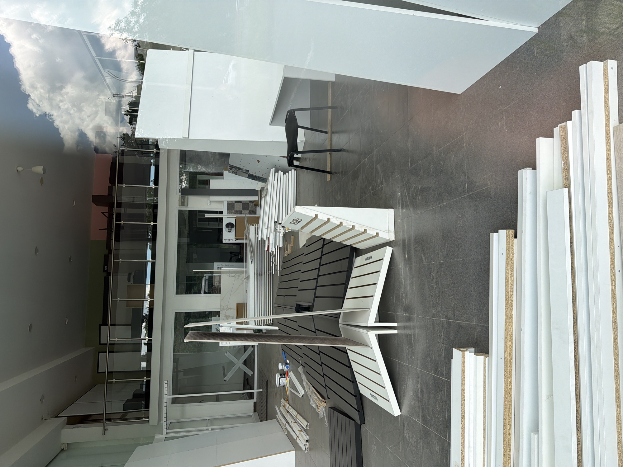 The image shows the interior of a shop, likely a showroom for furniture or interior design materials. Various display units and materials are visible, including white shelving and stacks of boards. The shop has large windows, allowing the outside scene to be reflected in the glass. Reflected in the window are clouds and buildings. Based on the Paravents branding on a piece of furniture and a reverse reflection of a website, the location is likely Burgas, Bulgaria. No people are directly visible in the image.
