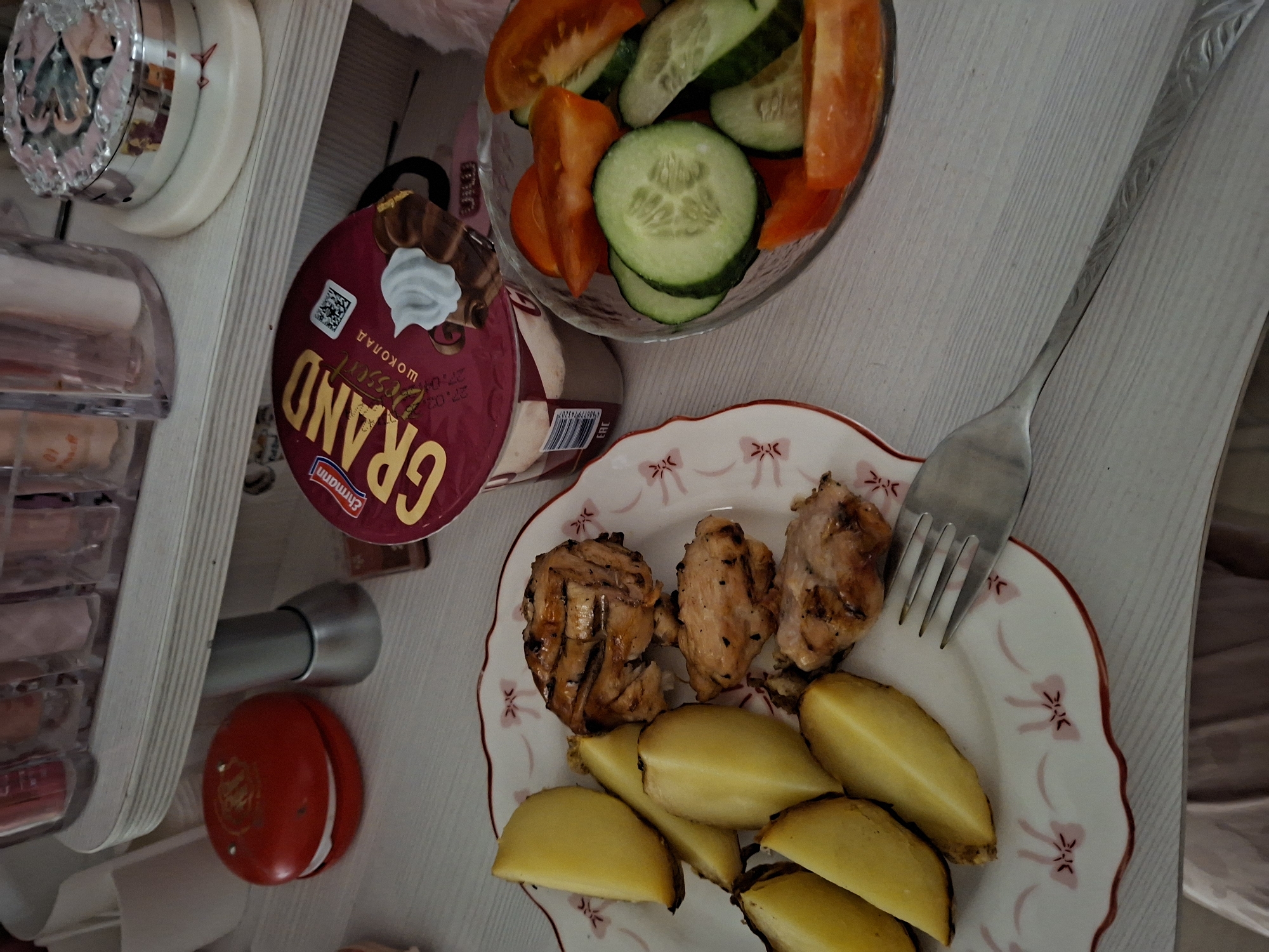 The image presents an overhead view of a meal and surrounding objects arranged on a light-colored, wood-grain tabletop in an indoor setting.
The main focus is a prepared meal. On a white plate with a red scalloped edge and a pattern of small pink bows, there are several pieces of grilled or pan-fried chicken with a browned, slightly charred appearance, alongside numerous golden-yellow wedges of roasted or boiled potatoes. A silver-colored fork with an embossed design on its handle rests on this plate.
Beside the main dish, a clear glass bowl contains a salad of sliced cucumbers and wedge-cut tomatoes, some red and some orange. To the left of the food items is a dark red plastic cup labeled "GRAND" in large yellow letters and "шоколад" (chocolate) in Cyrillic, with the brand "Ehrmann" also visible. The cup features a whipped cream graphic and a QR code, along with the date "27.03" printed twice, likely indicating an expiry date.
Further to the left, a clear, multi-tiered organizer, possibly for cosmetics, is visible. On top of it sits an ornate, reflective circular container. Below the organizer, a small red spherical object with unreadable gold lettering, a metallic cylindrical object, and a white cylindrical item are also present. The Cyrillic text on the dessert cup serves as a location cue, consistent with the provided context of Chaikovsky, Russia. The image captures a static moment of a meal setup.