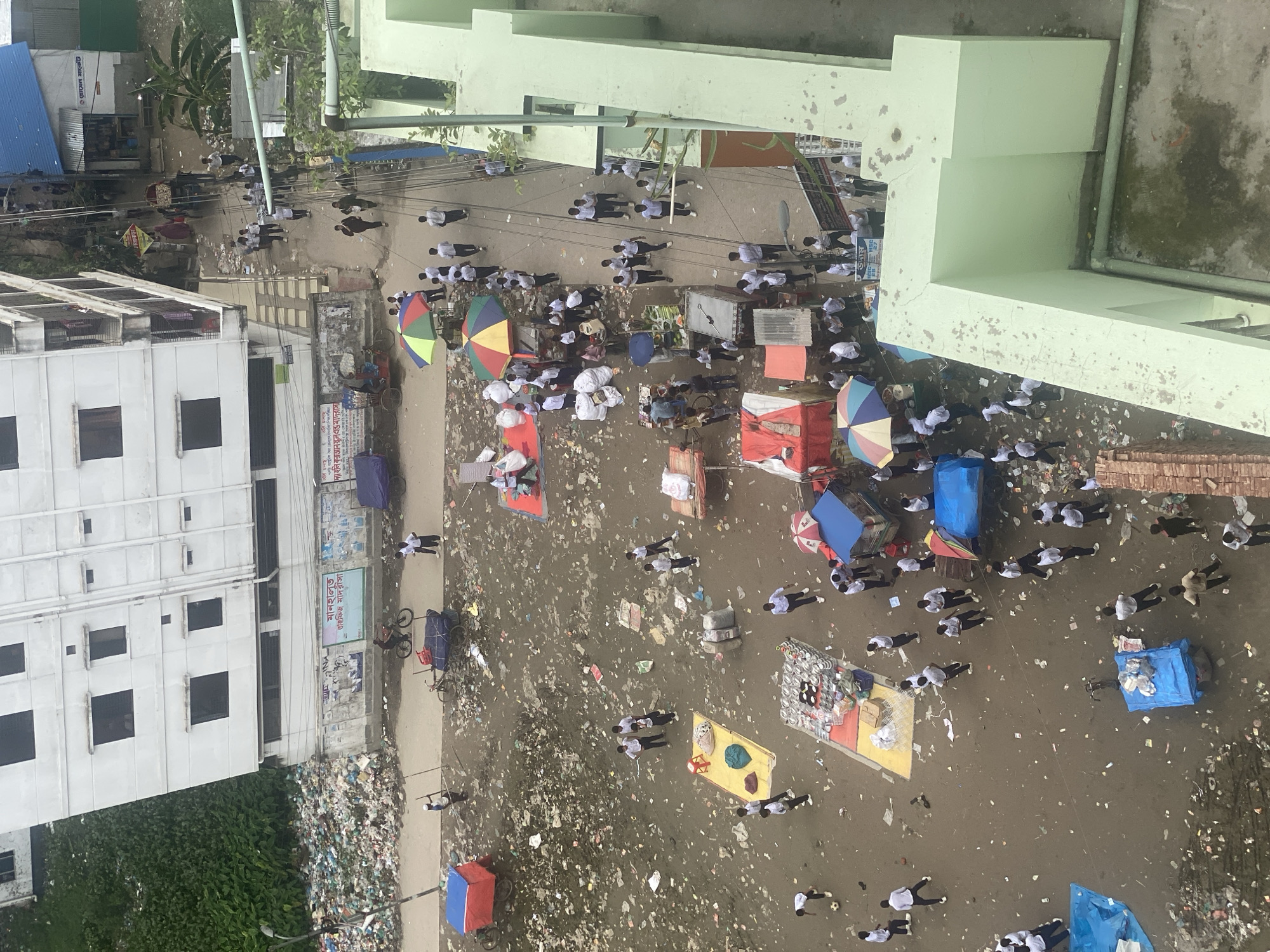 The aerial view shows a busy street scene in Dhaka, Bangladesh, characterized by a significant amount of scattered garbage and debris. Numerous people, many appearing to be students in white shirts and dark pants, are present on the street. They are seen walking individually and in groups, some gathered around market stalls, while others are engaged in informal activities like playing soccer. Several market stalls and carts are visible, displaying goods and providing shade with colorful umbrellas. The background includes buildings of varying heights, one of which is a white, multi-story structure. The environment is clearly cluttered with plastic waste, bags, and other refuse, suggesting a challenging urban sanitation situation. The overall impression is one of a vibrant but heavily polluted urban environment.
