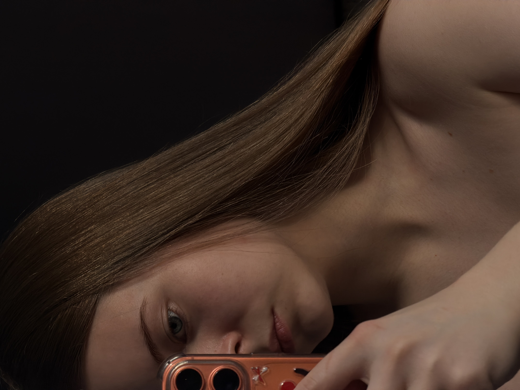 A young woman is taking a selfie in a dimly lit environment. She holds a smartphone with a transparent orange case. The background is dark, possibly black. The location is unknown and unrelated to Perm, Russia.
