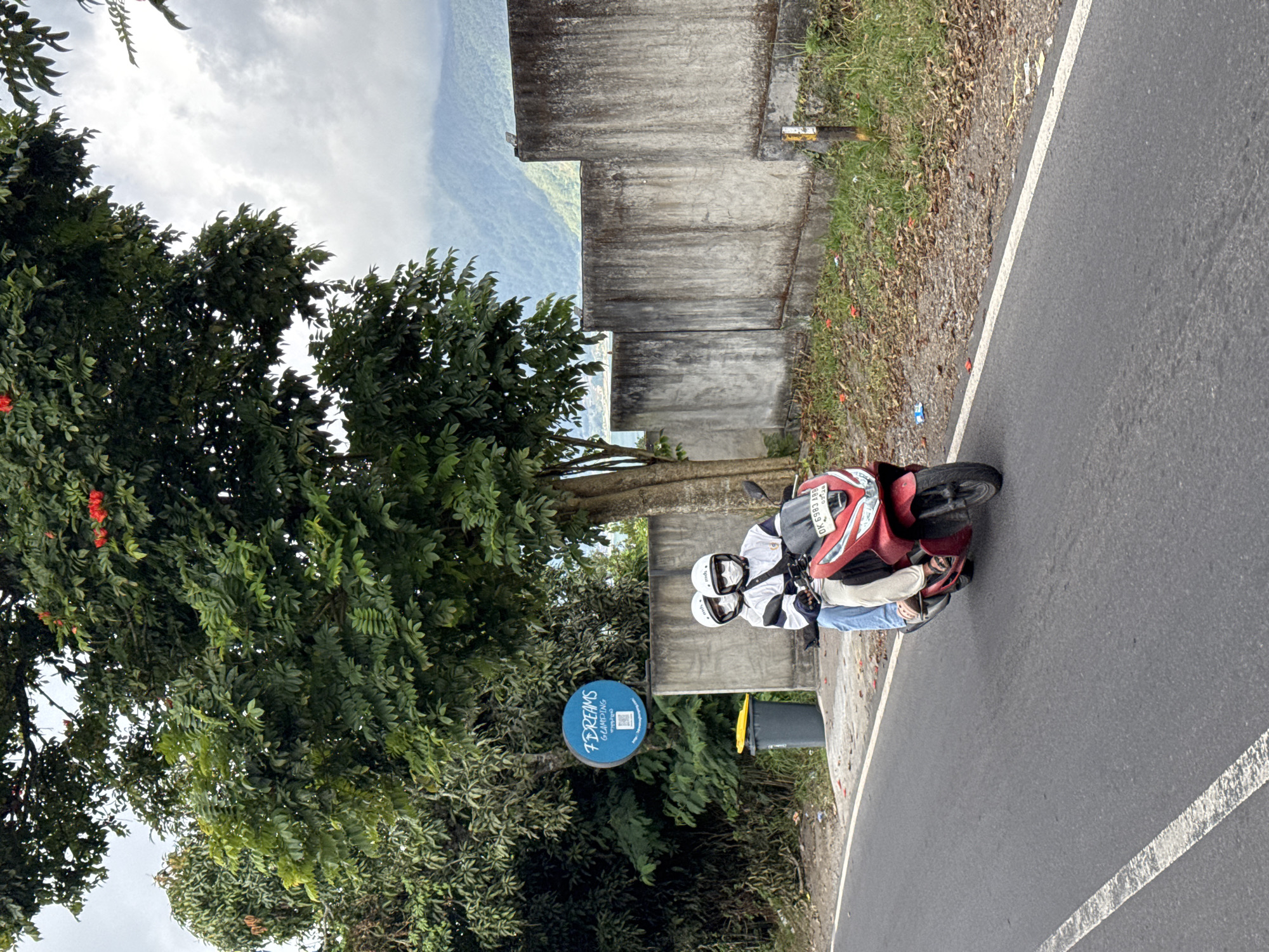 In Kintamani, Indonesia, two individuals wearing helmets and face masks ride a red scooter along a paved road. The rider in front wears a white jacket with black and white stripes, and the passenger behind wears a white jacket. They are positioned in the middle of the road, with trees and foliage to their left, and a concrete wall with vegetation and a post to their right. In the background, mountains are visible under a cloudy sky. A blue sign with white lettering, reading "7 DREAMS GLAMPING," is mounted on a pole near the road. A grey trash can with a yellow lid is situated next to the sign. The overall scene depicts a casual moment of travel on a scooter in a scenic, possibly rural, area.
