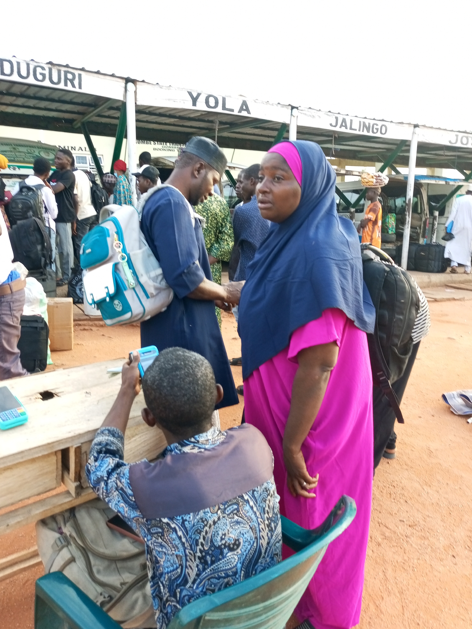 An outdoor scene at a bus terminal depicts a group of people standing and waiting. A woman wearing a bright pink dress and a dark blue hijab is in the foreground, looking to her right. Next to her, a man in a dark blue robe and a dark cap is looking down. In front of them, a young boy, wearing a patterned blue and white shirt, is seated and holding a small blue device, possibly a phone, in his hand. He appears to be looking at the device. Behind the woman, another person is visible with a backpack. In the background, there are signs with city names, including "YOLA", "JALINGO", and "JOS", suggesting a transportation hub. Other individuals and vehicles are present, and the ground is covered in sand or dirt. The lighting suggests it is daytime.