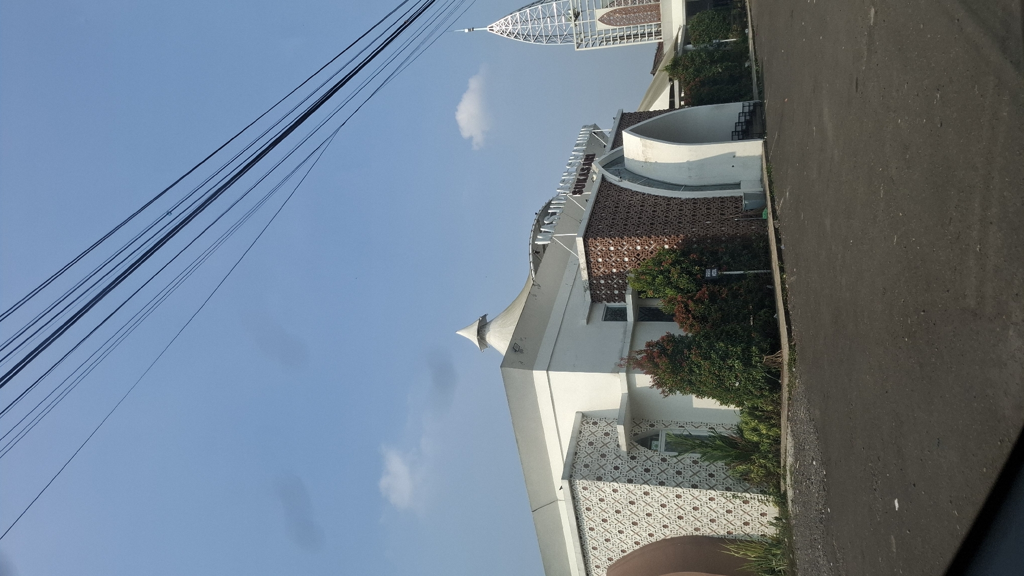 The image captures the exterior of a mosque in Sukasari, Indonesia, under a clear blue sky with scattered clouds. The mosque features a white, modern architectural design with prominent patterned panels in shades of brown and white adorning its facade. A distinctive conical roof caps the main structure, while a tall, slender, metallic spire rises to the right, also featuring patterned elements. Green foliage, including trees and shrubs, is present in front of the building, adding a touch of nature to the scene. An asphalt-paved area, likely a parking lot or walkway, occupies the foreground. Overhead, a series of power lines crisscross the frame against the sky. The text "MASJID" and "SUKASARI" is visible on the building's exterior, confirming the location.
