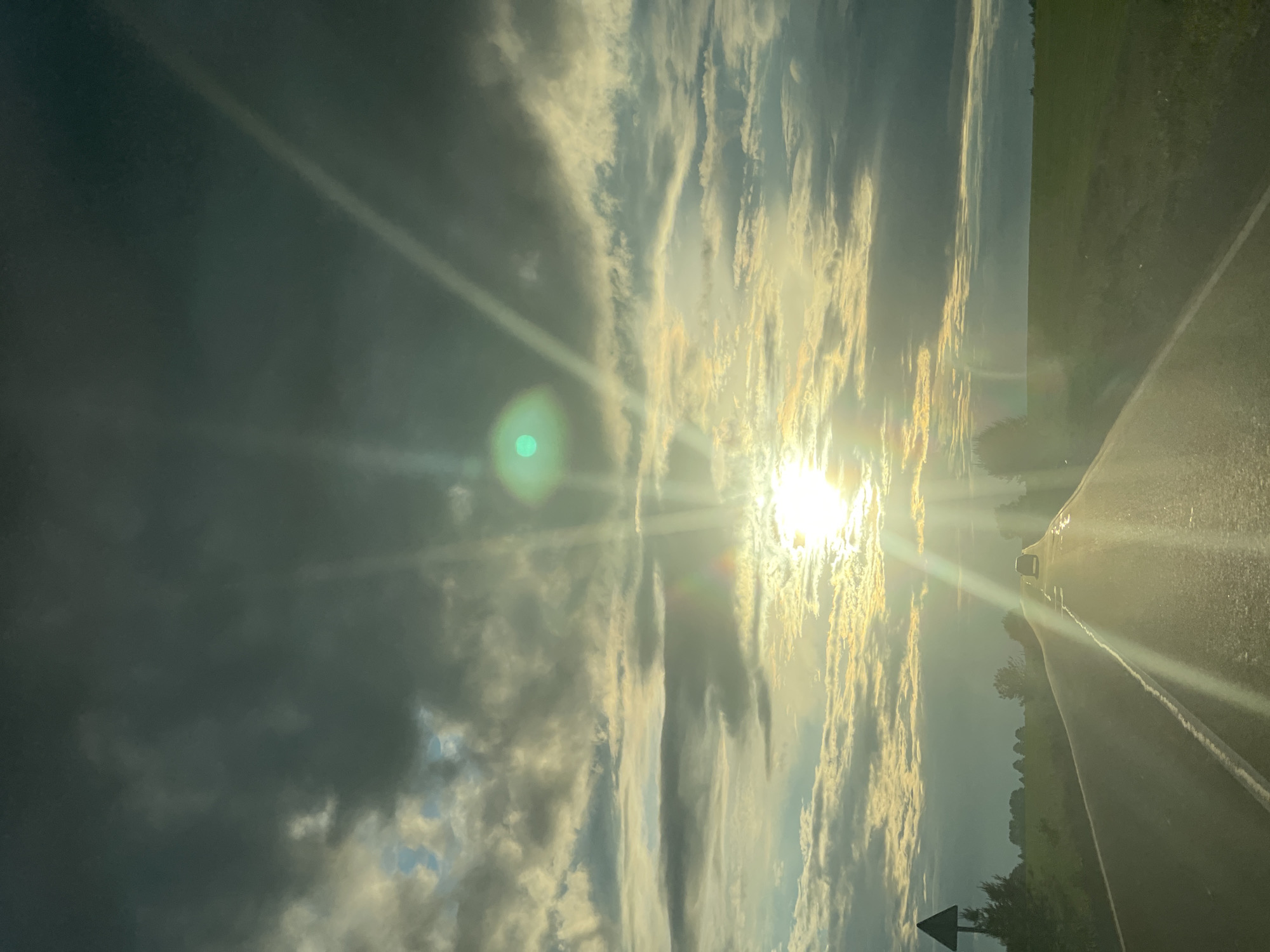 The image, when viewed in its correct orientation, captures a dramatic sky at what appears to be either sunrise or sunset. A powerful, brilliant sun dominates the right side of the frame, emitting intense white and golden light that creates prominent sunbeams and a noticeable green lens flare in the upper left.
The sky is a dynamic canvas of clouds; darker, heavier formations occupy the left side, transitioning to lighter, elongated clouds that are brilliantly illuminated by the low sun on the right, glowing in shades of yellow and orange.
Below the radiant sky, a road extends diagonally from the lower right foreground into the distance. It is an asphalt surface with a visible white edge line, reflecting the strong sunlight. A dark vehicle is partially visible on the road in the mid-ground, silhouetted by the intense backlighting. To the left of the road, a dark landscape of trees and fields stretches out, also appearing largely as silhouettes against the brighter sky. A dark, triangular road sign with a white arrow pointing to the right is visible in the lower left corner. The scene evokes a sense of journey during the golden hour, marked by striking natural light and cloud formations. No visible text is present.