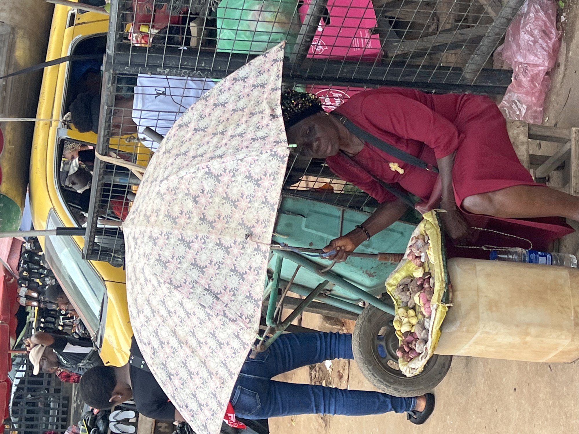 A street scene in Nkpor, Nigeria, features an outdoor market. In the foreground, a woman sits on a wooden stool under a floral umbrella, wearing a red dress and a beaded headscarf. In front of her, a tray of various tubers, possibly yams or cassava, rests on a tan plastic container. Her hand is visible reaching towards the tray. To her left, a young person in blue jeans and a black shirt stands. Behind them, a yellow vehicle is parked, with people visible inside. A metal fence encloses part of the scene, and various market stalls and goods, including bags and footwear, are visible in the background. The ground appears to be dirt or rough pavement.