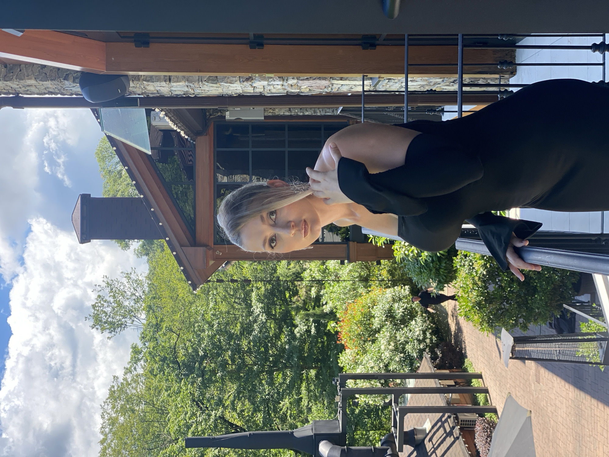 The media file shows a young woman in a black dress, leaning against a railing. She has blonde hair and is looking at the camera with a neutral expression. The background is a blur of green trees and a brick pathway. In the distance, another person in black is walking away from the camera. The scene appears to be outdoors, possibly on a balcony or patio, with a building structure visible in the background. The lighting suggests it is daytime, and the presence of trees and greenery indicates a warm climate. The sky has some clouds. The overall impression is a casual portrait taken in an outdoor setting.