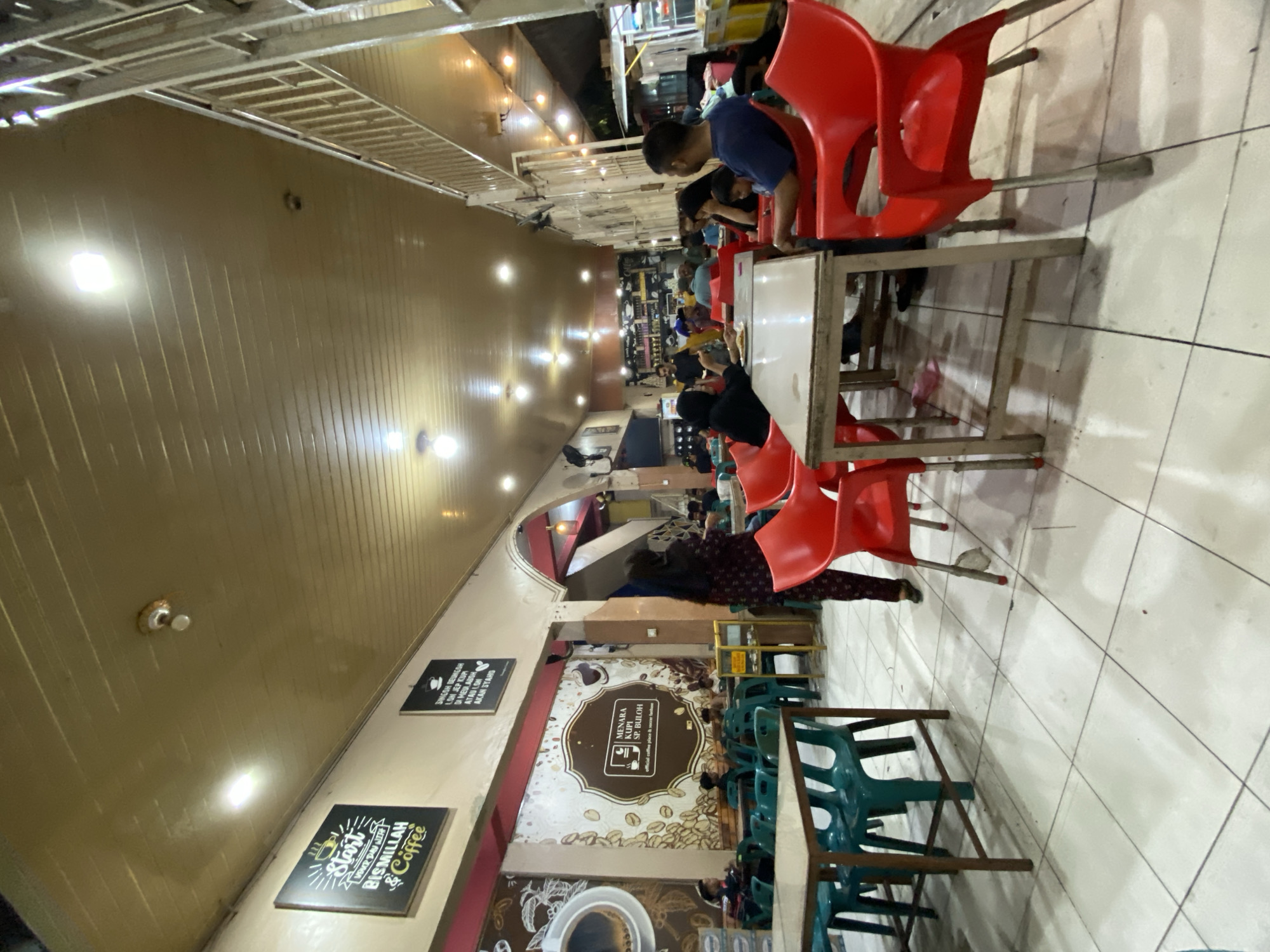 This image depicts the interior of a cafe in Lhokseumawe, Indonesia, likely during the evening given the artificial lighting. The scene is filled with people seated at tables, engaged in various activities, possibly dining or socializing. Red plastic chairs are prominently featured, surrounding wooden tables, with some green plastic chairs also visible.
The cafe appears to have a simple, functional design with a light-colored, paneled ceiling and tiled floors. Several signs with text are visible on the walls. One sign, in black and white, reads "Start Your Day with Bismillah & Coffee," suggesting a focus on coffee and perhaps a religious or cultural invocation. Another sign, also in black and white, features text in a language that appears to be Bahasa Indonesia, possibly related to the cafe's offerings or a motto. A larger, circular graphic with coffee beans and text reading "MENARA KUPI SP. BULOH" and "Official coffee place & secret recipes" clearly identifies the establishment as "Menara Kupi Sp. Buloh."
The lighting is somewhat dim, with bright spotlights creating illumination, and the overall atmosphere seems relaxed and casual. The individuals in the image are mostly seen from behind or in profile, making it difficult to discern specific emotions, but the presence of multiple patrons suggests a moderately busy environment. The perspective of the photo is from a slightly elevated angle, looking down into the cafe space.