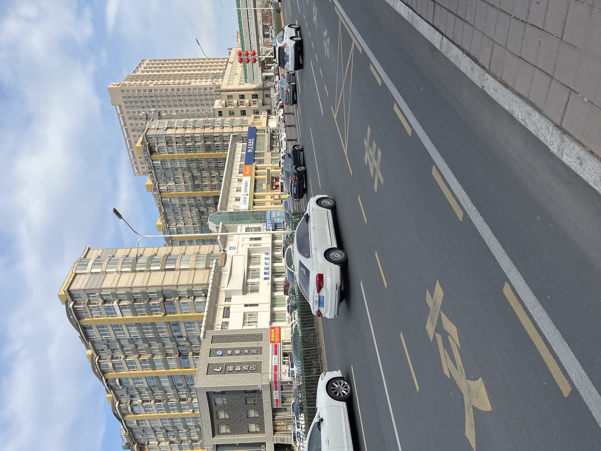 This image presents a street scene in Changchun City, China, lined with tall buildings and showing vehicles on a gray asphalt road. Several cars, mostly white, are visible on the street, either parked or driving. Building facades display various signs and logos in Chinese, including "JLRCB," "麗枫酒店," and "吉优客." Yellow markings are painted on the road surface, indicating traffic lanes and potentially prohibited parking areas. A traffic light is visible in the distance, and the sky above is a mix of blue and clouds. The buildings appear to be mixed-use, with commercial spaces at street level and residential or office spaces above. The photograph is taken from a slightly elevated perspective, looking down the street.