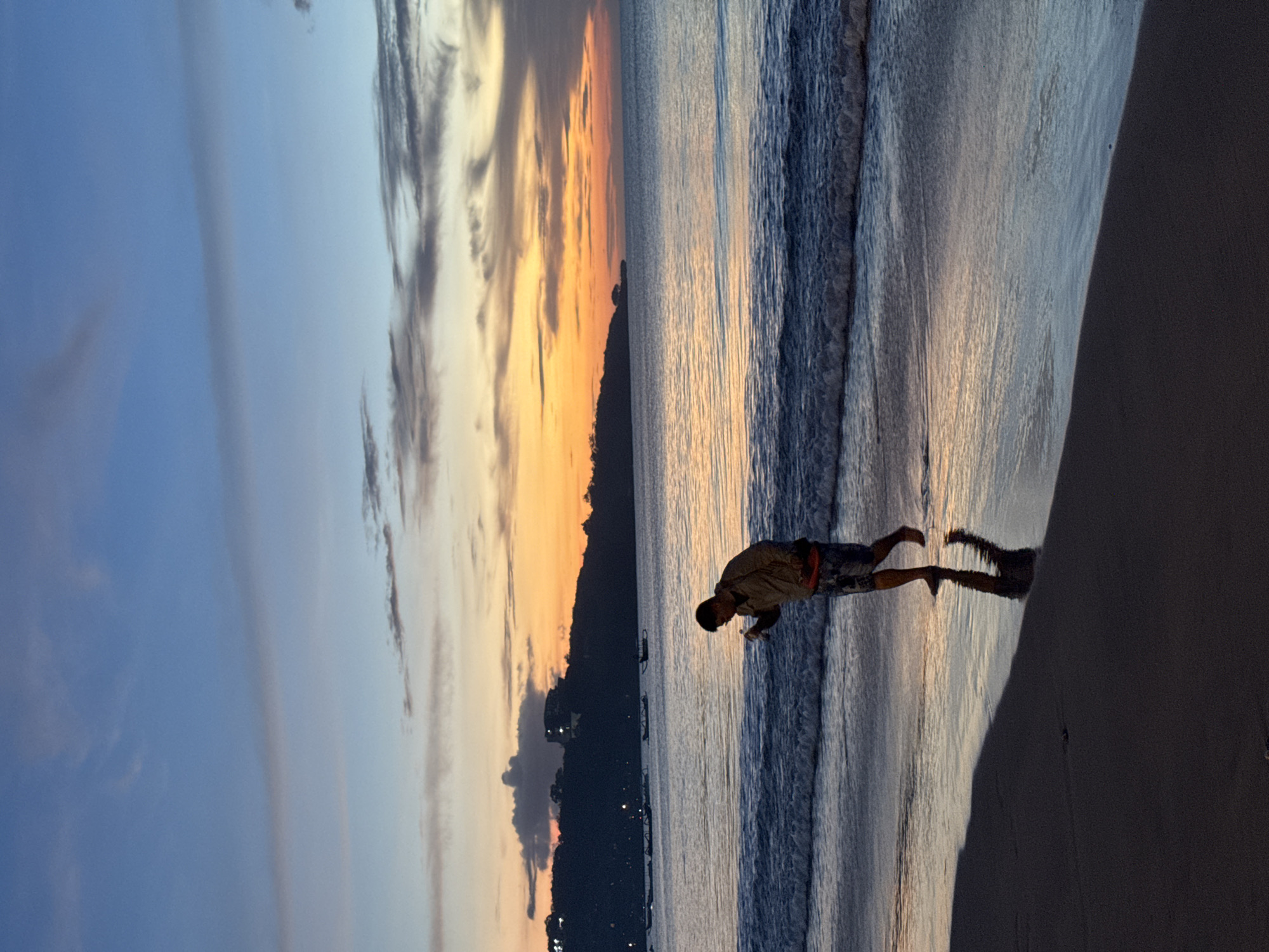 This landscape image captures a serene beach scene in Kuta Selatan, Indonesia, during either sunrise or sunset. The sky, covering the upper left, displays a gradient of soft blue, blending into warm oranges and yellows closer to the horizon on the right. Wispy, dark clouds are interspersed across the colorful sky, particularly concentrated above the distant landmass.
A silhouetted mountain range or coastline stretches across the horizon, appearing dark against the luminous sky. Scattered lights, indicative of a town or settlement, twinkle faintly along the base of this landform. Several small boats are faintly visible near the distant shore.
The ocean dominates the middle of the frame, its surface gently rippled and reflecting the vibrant colors of the sky. Small waves are visible breaking onto the shore, creating a foamy edge. A lone individual, appearing to be a male, stands in the shallow water near the sandy beach in the mid-right portion of the image. He is dressed in a light-colored shirt and patterned shorts, leaning slightly forward as if observing something in the water or at his feet. His silhouette is cast long across the wet sand and water, complementing the mood of the scene.
The sandy beach occupies the lower right, dark and wet from the receding tide. The overall impression is one of tranquility and natural beauty, highlighting a peaceful moment by the sea during the golden hour. No visible text is present.