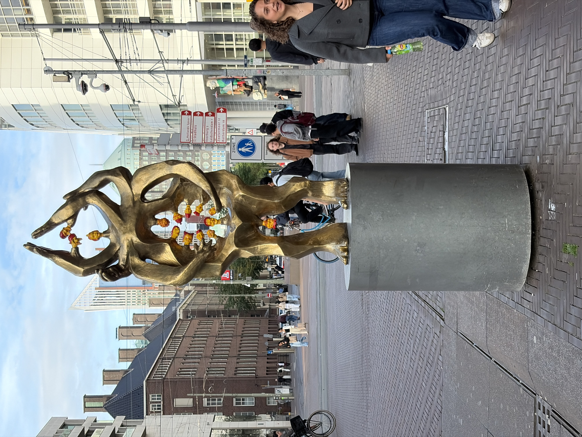 This image depicts a detailed urban street scene in The Hague, Netherlands. The specific location is near The Hague City Hall (Stadhuis) and the Muzentoren skyscraper, identifiable by architectural features and street signage.
In the center foreground stands a large bronze sculpture, approximately 2.5 meters tall, resting on a cylindrical grey concrete or stone pedestal about 1 meter high and 1 meter in diameter. The sculpture consists of several intertwined, abstract human-like figures. Within openings in the sculpture's structure, numerous smaller, brightly colored elements resembling stylized human heads or fish, primarily in shades of yellow, orange, and red, are suspended.
To the immediate right of the sculpture, a young adult female with curly brown hair, wearing a dark grey jacket, dark blue jeans, and white sneakers, walks from right to left. She holds a green Lipton iced tea bottle in her left hand and smiles slightly.
In the mid-ground, behind the sculpture, several pedestrians are visible. A man in a brown jacket stands talking to another individual wearing a grey hoodie and a red backpack. Other people walk or ride bicycles in various directions across the brick and paved street.
The background features a mix of architectural styles. To the left, older, multi-story brick buildings with dark tiled roofs and multiple chimneys are present. Further left, a modern, tall skyscraper with a distinctive triangular peak (the Muzentoren) rises above the older buildings. Another building with a green domed or pyramid-shaped roof is visible in the distant left. To the right, a prominent, large, multi-story white building with numerous windows, identified as The Hague City Hall, dominates the skyline.
Street furniture includes a red and white street signpost indicating directions to "Hollands Spoor," "Stadhuis," "Voorburg," "Leidschendam," "Zoetermeer," and "Rotterdam." Below it is a blue "ZONE" sign with a pedestrian symbol. Multiple utility poles with various attachments, including two CCTV cameras, are visible on the right, along with overhead tram lines. The ground is paved with a combination of dark brick patterns (herringbone and linear) and lighter grey concrete slabs, which include drainage grates. A minimal patch of green vegetation is visible at the base of the sculpture's pedestal. The sky is partially cloudy, suggesting daylight conditions.