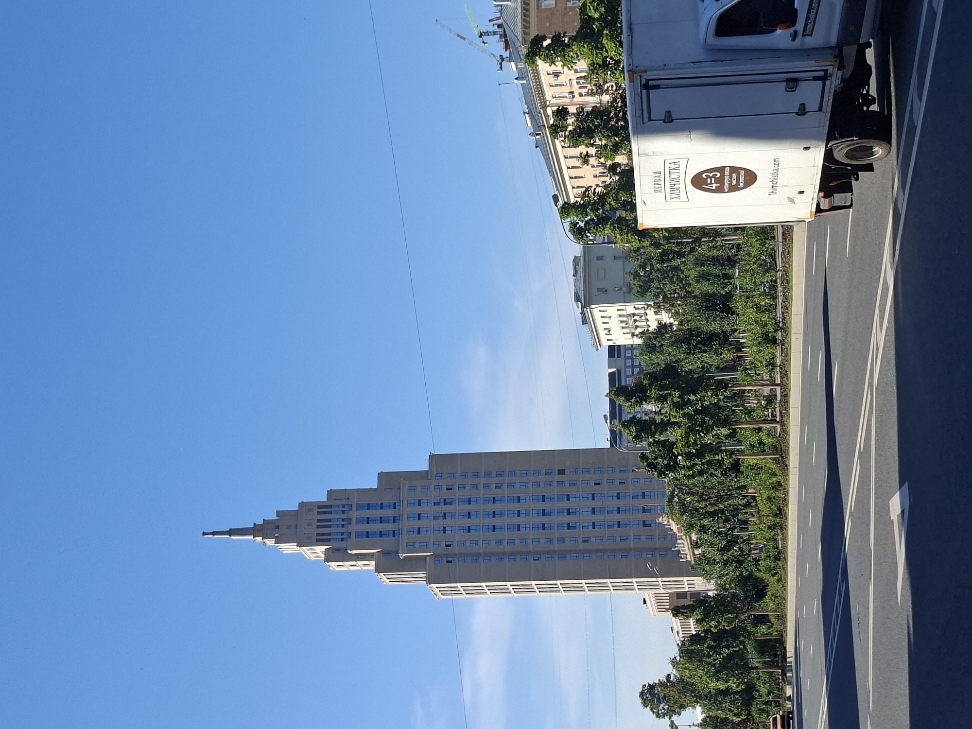 The image depicts a bright, sunny day in Moscow, Russia, dominated by a towering Stalinist skyscraper. The skyscraper, located at the intersection of Kutuzovsky Prospekt and Trekhgornaya Ulitsa, is a prominent landmark in the city. The building features a distinctive spire and numerous windows, suggesting it is either an apartment building or a government office. In the foreground, a white delivery truck with a Russian advertisement for a dry cleaner ("ПЕРВАЯ ХИМЧИСТКА") is partially visible, traveling on a multi-lane road. The road is marked with white lines and a directional arrow, indicating traffic flow. Lush green trees line the side of the road, with other buildings and construction cranes visible in the background, hinting at ongoing development in the area. The sky is clear and blue, contributing to the overall sense of a pleasant day.