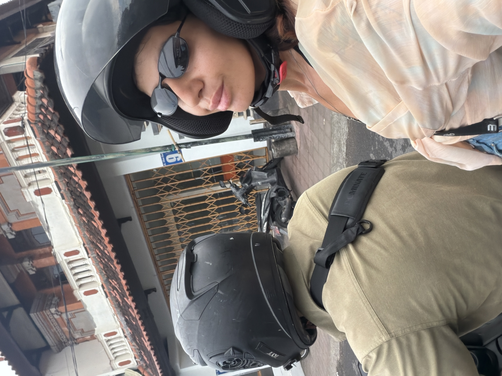 The image captures a close-up view of two individuals on a motorcycle in Kuta Selatan, Indonesia. The person in the foreground, a woman with dark hair, wears a black helmet and sunglasses. She is looking towards the camera, with her mouth slightly open. She is dressed in a light-colored, translucent blouse and a necklace. The person behind her, likely male based on the broad shoulders, is wearing a black helmet and a tan shirt. A black strap with white text ("NOMINATE") is visible across his back. The background shows a building with a tiled roof and ornate architectural details, characteristic of Balinese style. A metal grate is visible on the building's lower level, and a street sign with the number '9' is mounted on a pole. A parked scooter is visible in the mid-ground. The overall scene suggests a casual ride through a bustling urban or semi-urban environment in Kuta Selatan.