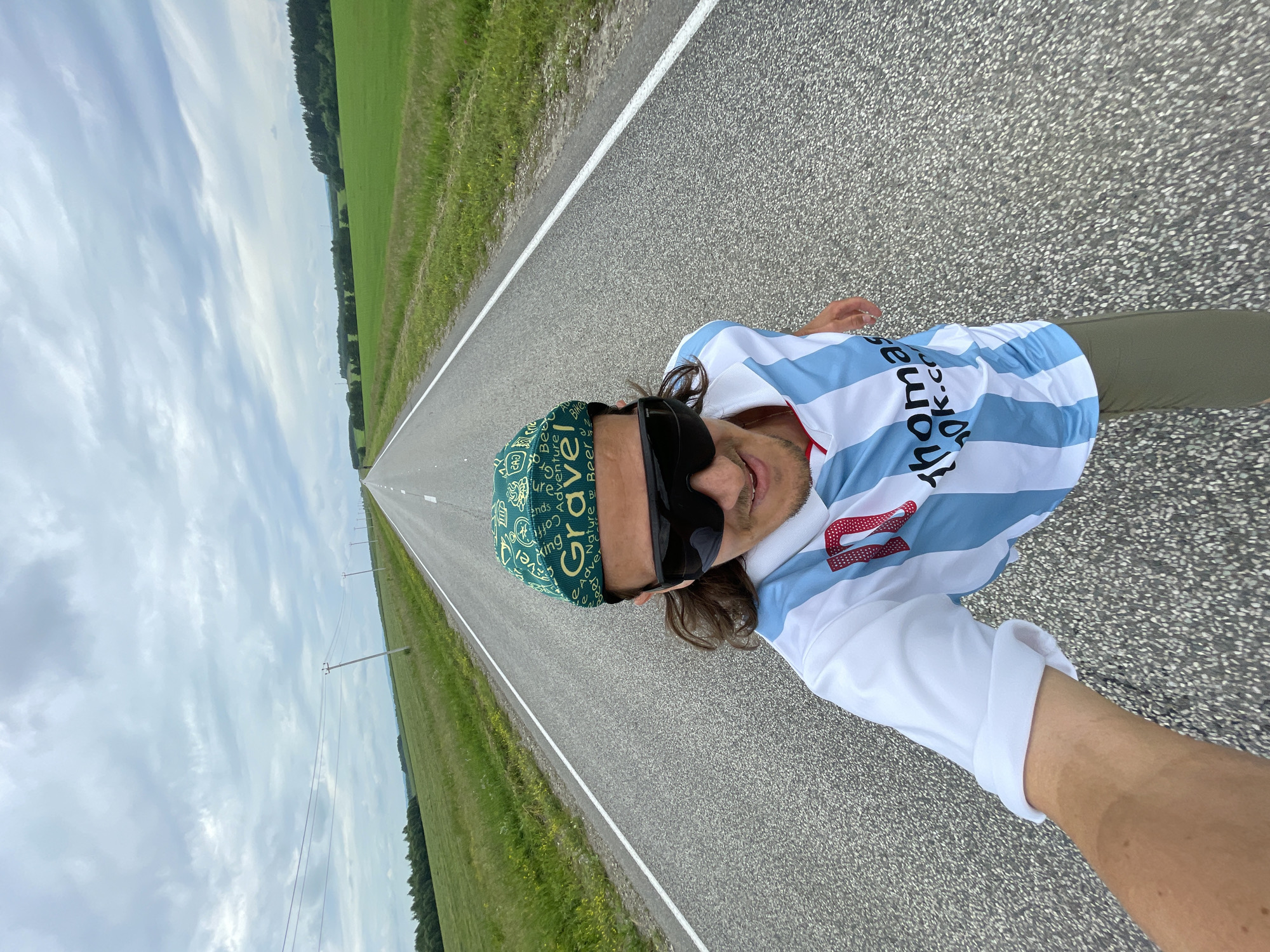 A man is captured in a wide-angle, front-facing selfie as he stands on an empty asphalt road stretching into the distance. The road is bordered by vast, green fields on either side, with scattered trees and power lines visible in the background under an overcast sky. The man is wearing a green bandana with white lettering, large black sunglasses, and a white and light blue striped sports jersey with a red number '10' on the chest. He is also wearing olive-colored pants. The image appears to have been taken outdoors in a rural setting.