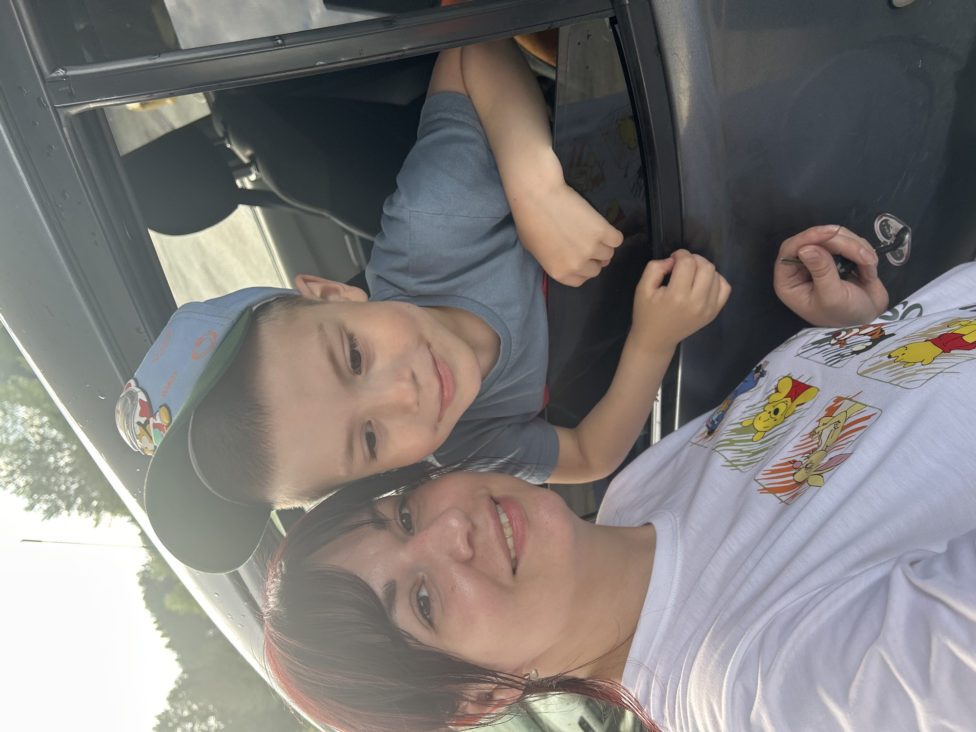 The image shows a woman and a young boy in a car. The woman is in the foreground, looking at the camera and smiling. She has dark hair and is wearing a white t-shirt with cartoon characters printed on it. The boy is in the background, leaning out of the car window. He has short hair and is wearing a blue t-shirt and a baseball cap. The car appears to be parked on a street in Chaikovsky, Russia, with trees visible in the background. The lighting suggests it is daytime.