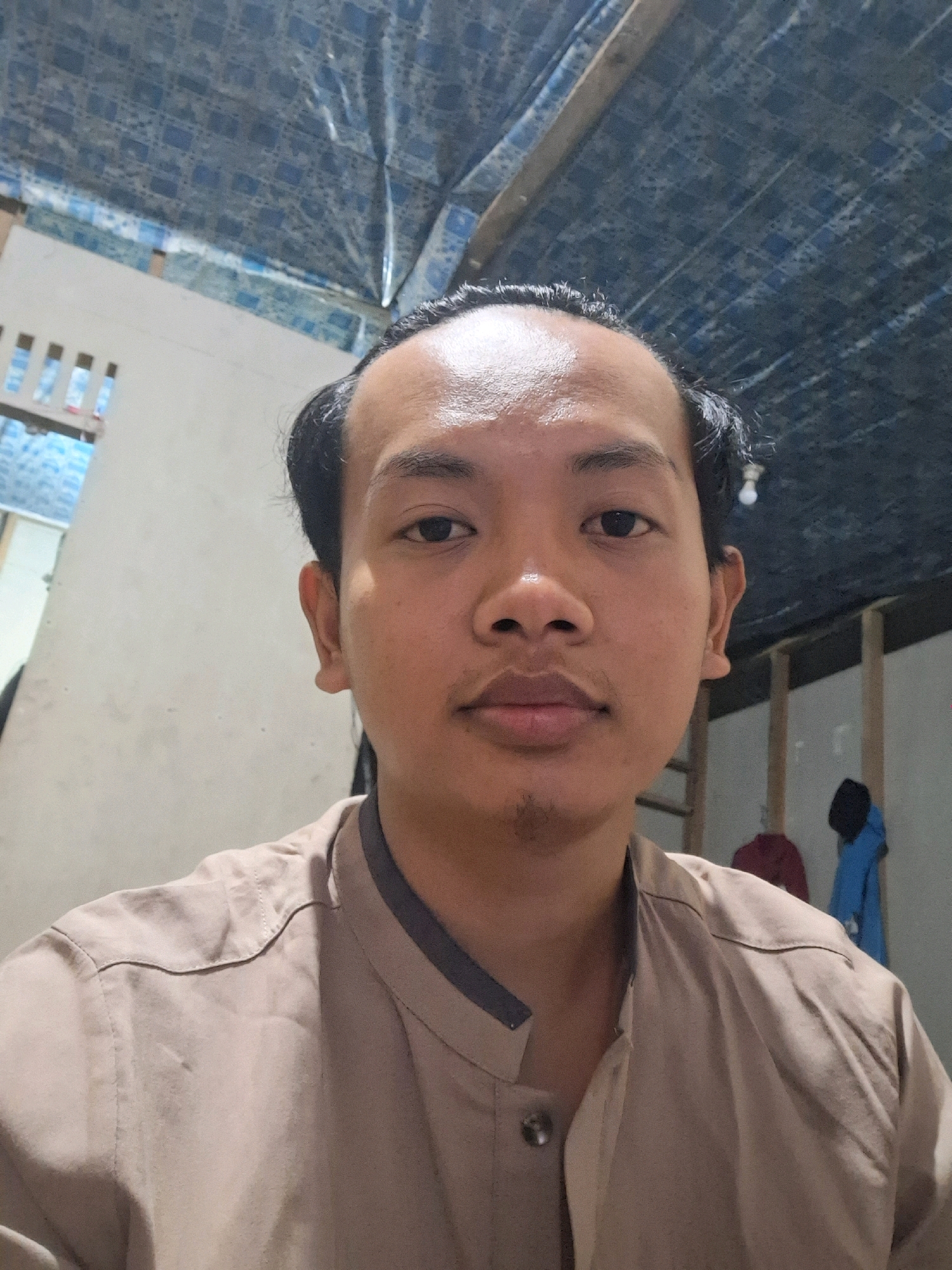 A medium shot is captured of a young man posing for a selfie. He is light-skinned with dark hair, which is slicked back from his forehead. His facial features include dark eyes, a prominent nose, and full lips. He has a faint mustache and goatee. He is wearing a beige button-up shirt with a dark grey collar. The background suggests an indoor setting with a corrugated plastic roof, possibly in a room with white walls and wooden structural elements. On the wall to the right, two garments, one red and one blue, are hanging. To the left, a white wall with a slatted window is visible. The image appears to be taken in Air Tawar, Indonesia.