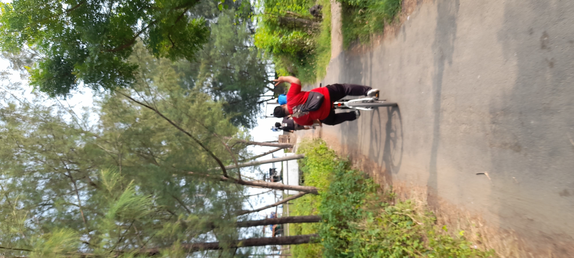 The image depicts a group of people cycling on a paved road lined with trees on both sides. In the foreground, a person wearing a red shirt and black pants is riding a bicycle, with a black bag slung over their shoulder. They are raising their right hand, displaying two fingers in a peace sign. Behind them, two other individuals on bicycles are visible, one in a black shirt and another in a blue shirt. The road appears to be in a rural or semi-rural setting, possibly near a beach, as indicated by the presence of palm trees and the bright sky. The overall scene suggests a recreational activity taking place in an outdoor environment. The location is Nyamplung Sari, Indonesia.