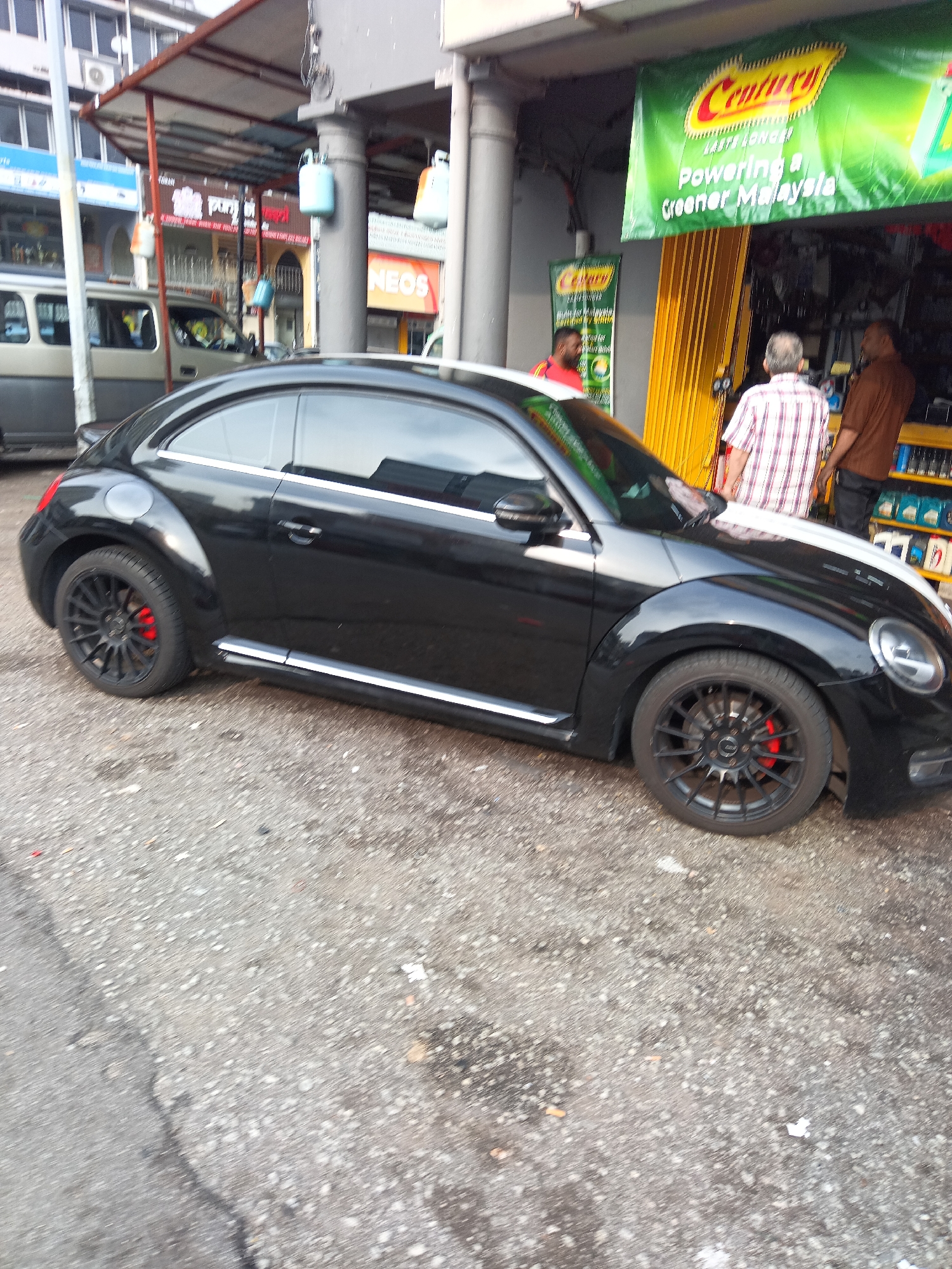 A black Volkswagen Beetle is parked on a gravelly street in front of a shop in Kuala Lumpur, Malaysia. The car has black alloy wheels and red brake calipers. Behind the car, a man wearing a checkered shirt stands next to another man wearing a brown shirt. A yellow retractable security gate is open, revealing shelves stocked with bottles and cans. A green banner with yellow lettering hangs above the entrance, displaying the word "Century" and the slogan "Powering a Greener Malaysia." In the background, a silver van is partially visible, parked in front of a building with multiple windows. The sky is overcast.